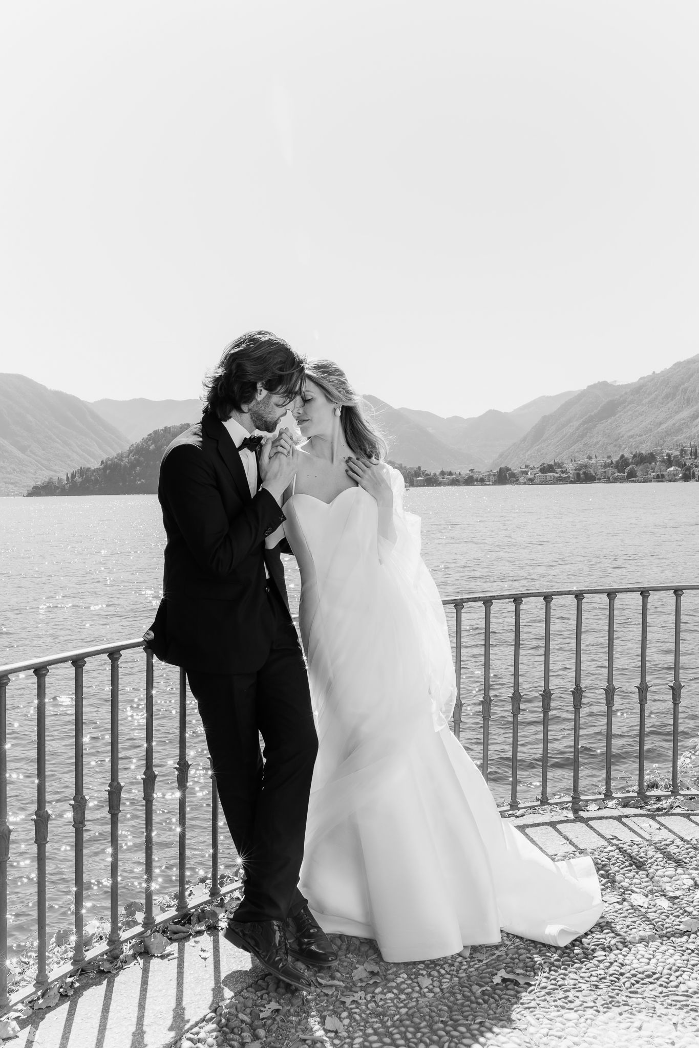 Black and white photo of a bride and groom sharing a romantic kiss by a lakeside railing with mountains in the background.