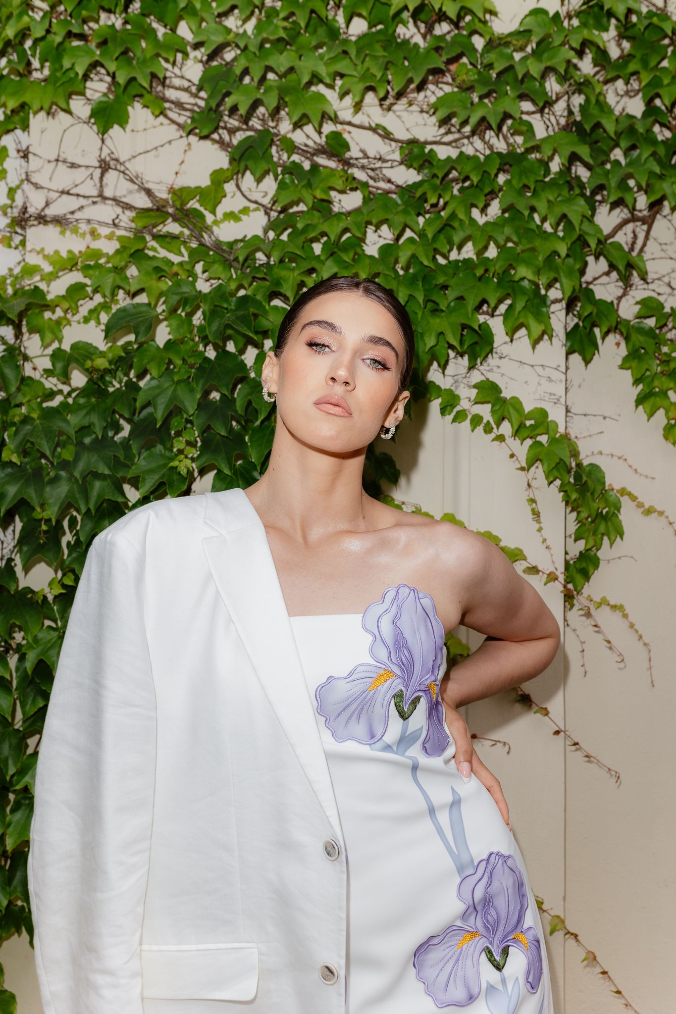 Woman in white blazer and floral iris dress poses against a wall with lush green ivy backdrop.
