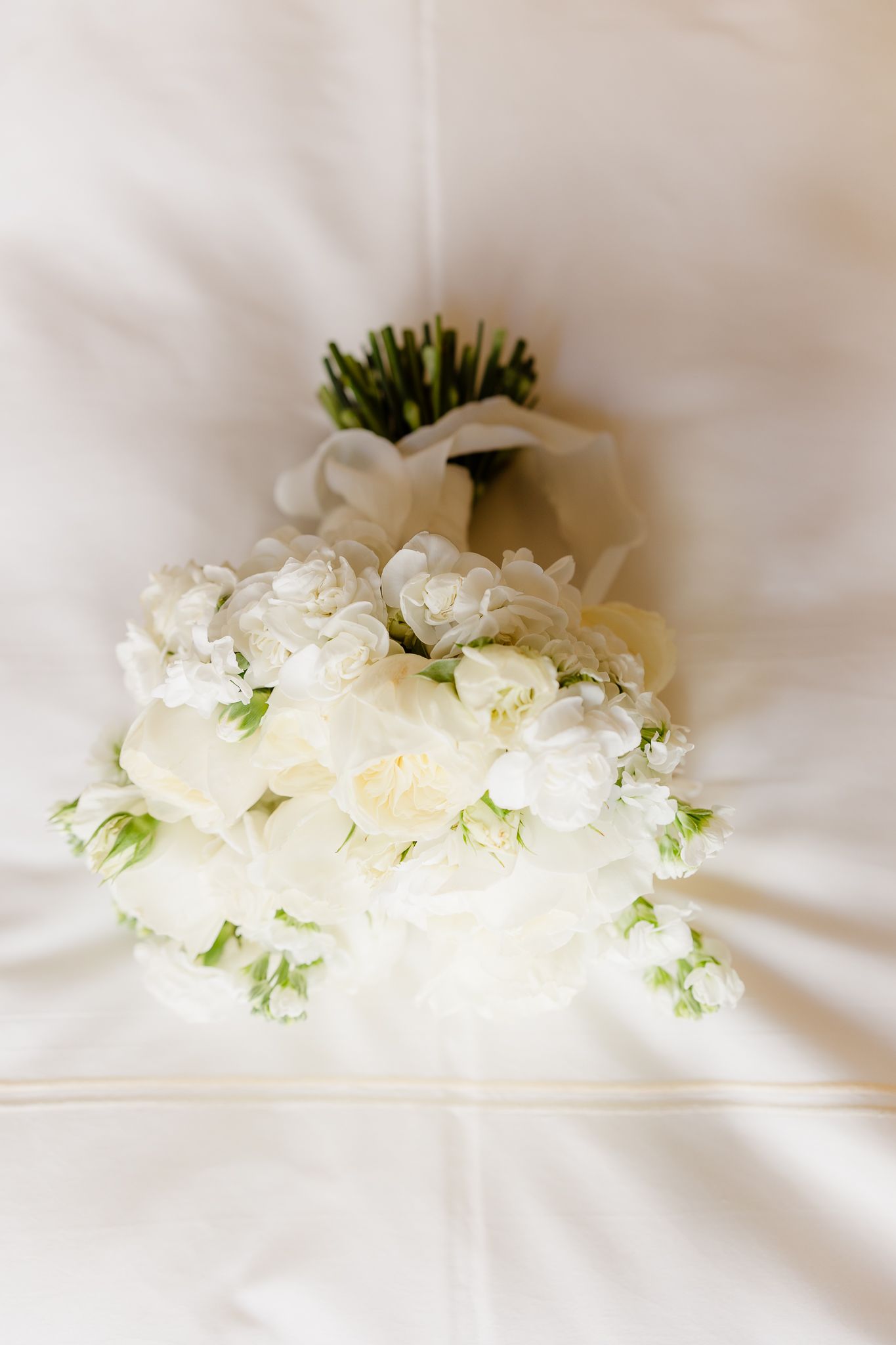 Classic all-white wedding bouquet of roses and small blooms tied with a white ribbon on white fabric.
