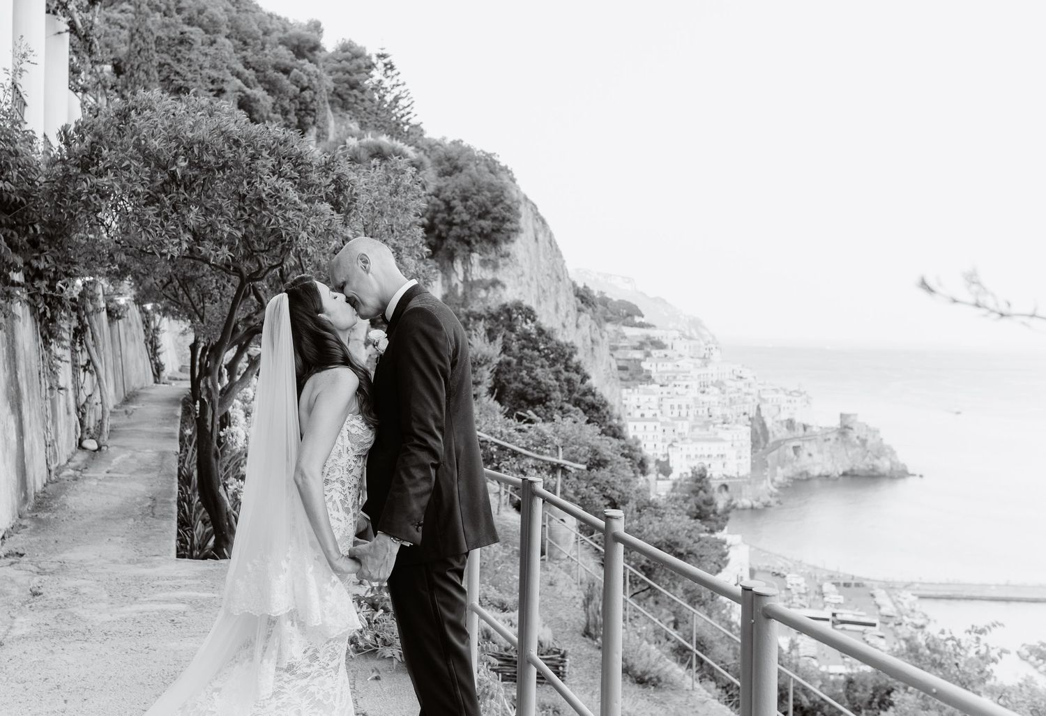 Black and white photo of bride and groom kissing on scenic Amalfi Coast overlook with ocean views.
