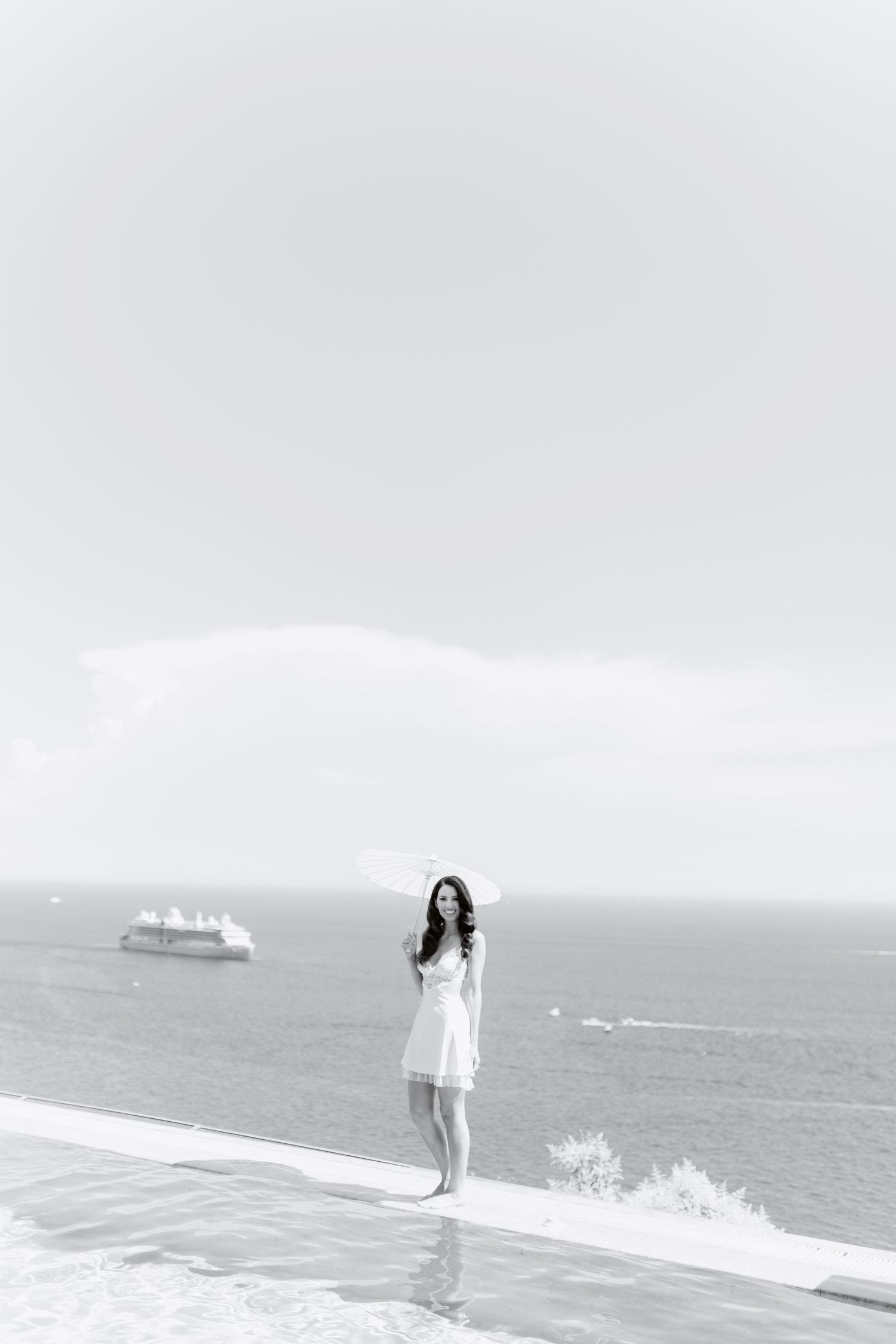 Black and white photo of a woman in a white dress standing by an infinity pool overlooking the ocean with a cruise ship.