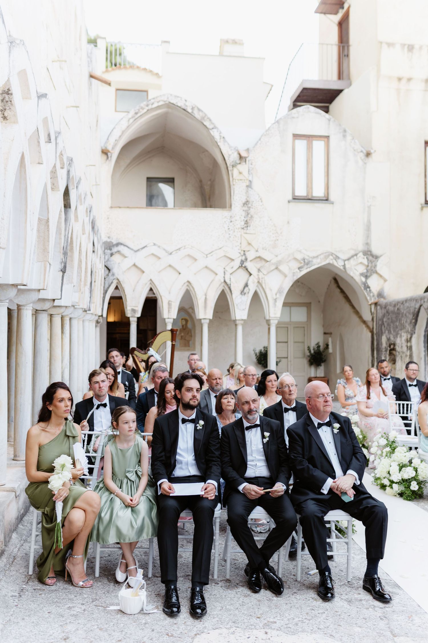 Guests seated at an outdoor wedding ceremony in a stunning white arched courtyard in Italy.
