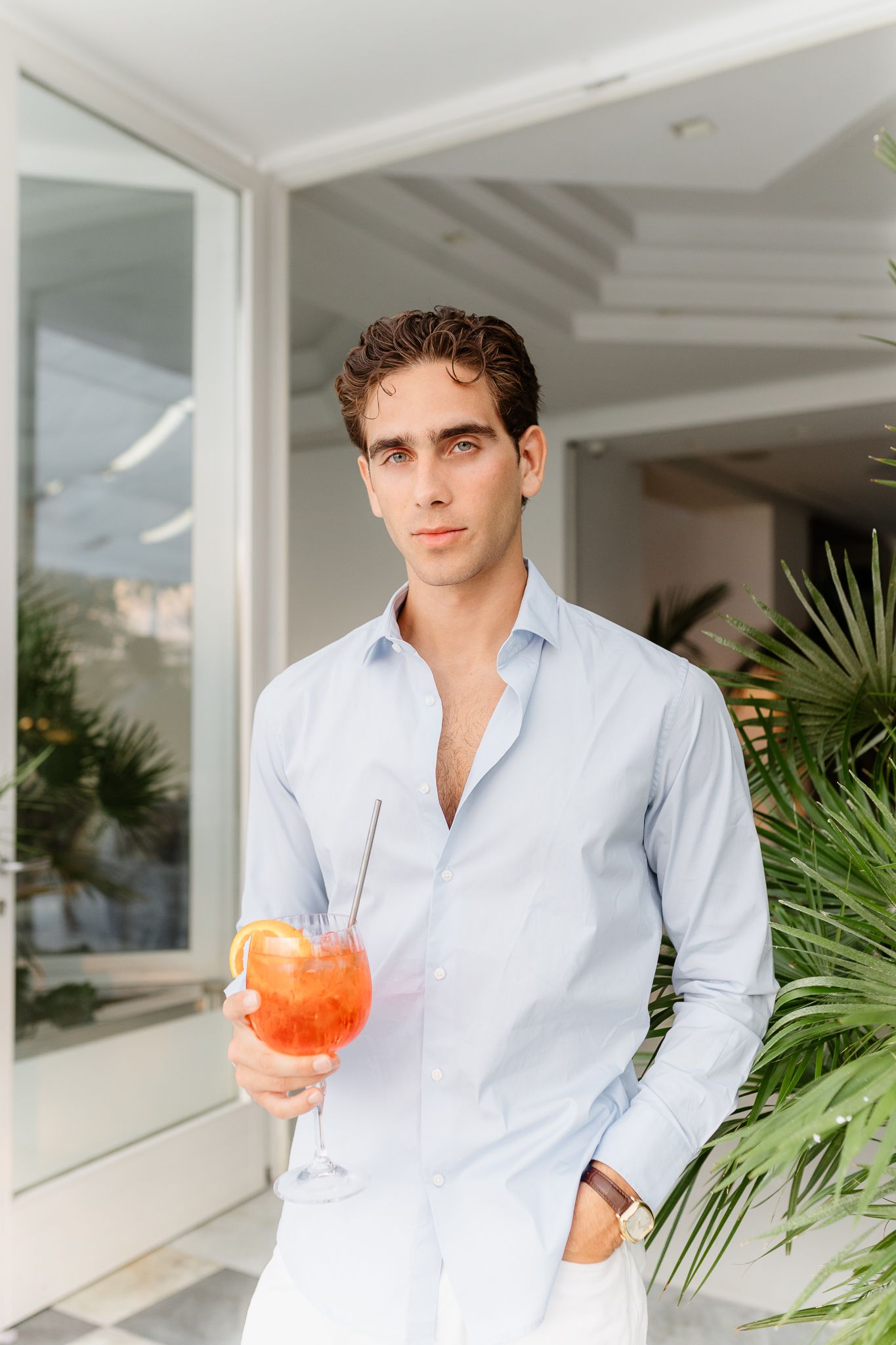Young man in light blue shirt holding an Aperol Spritz cocktail outdoors near tropical plants.
