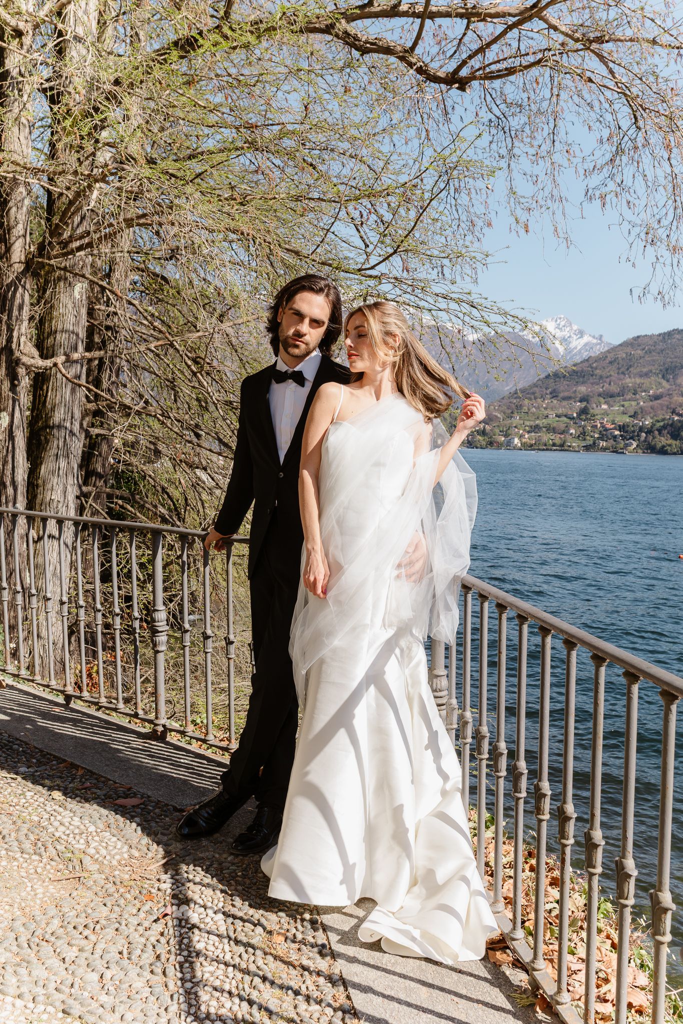 Bride and groom pose together on sunny lakeside promenade with snow-capped mountains in background.