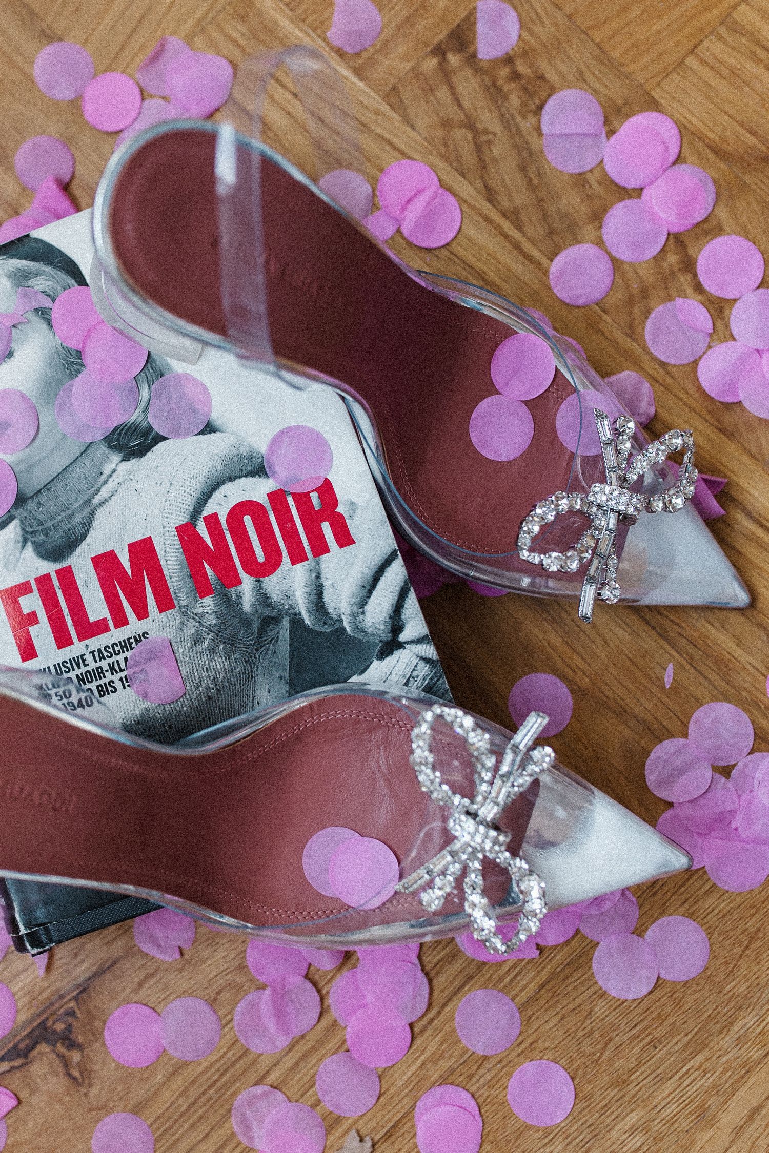 Crystal bow clear heels beside a Film Noir book surrounded by pink confetti.