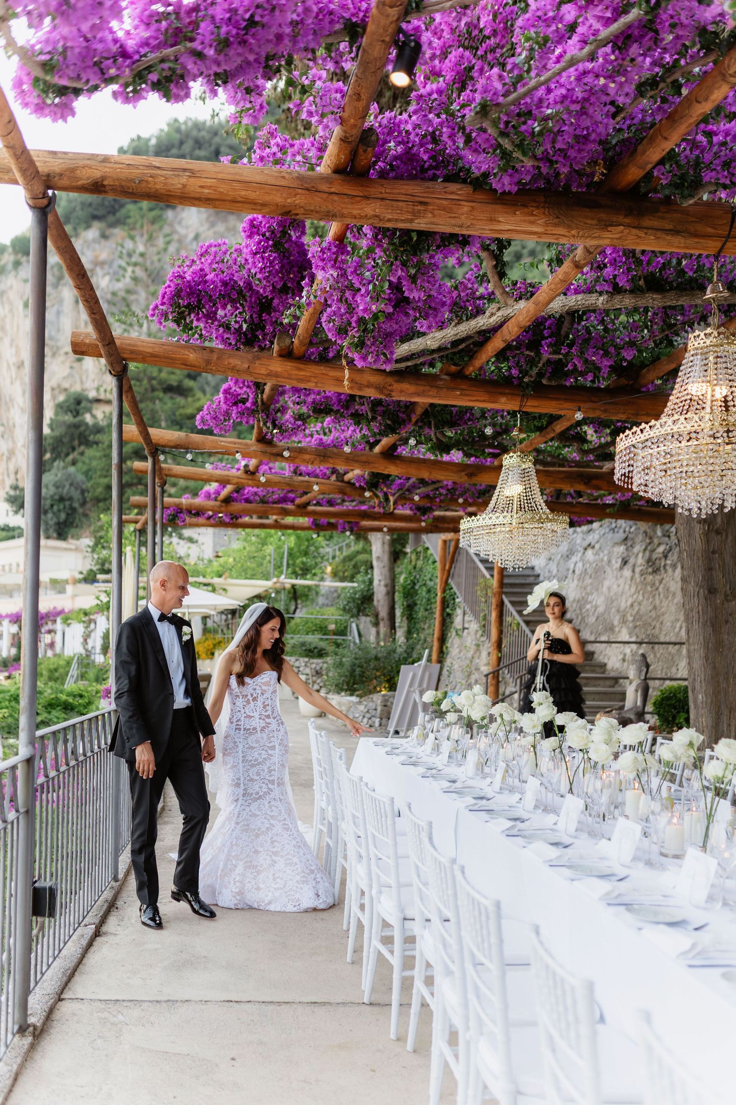 Bride and groom walk beneath a stunning purple bougainvillea pergola beside an elegant white reception table in Anantara Amalfi.