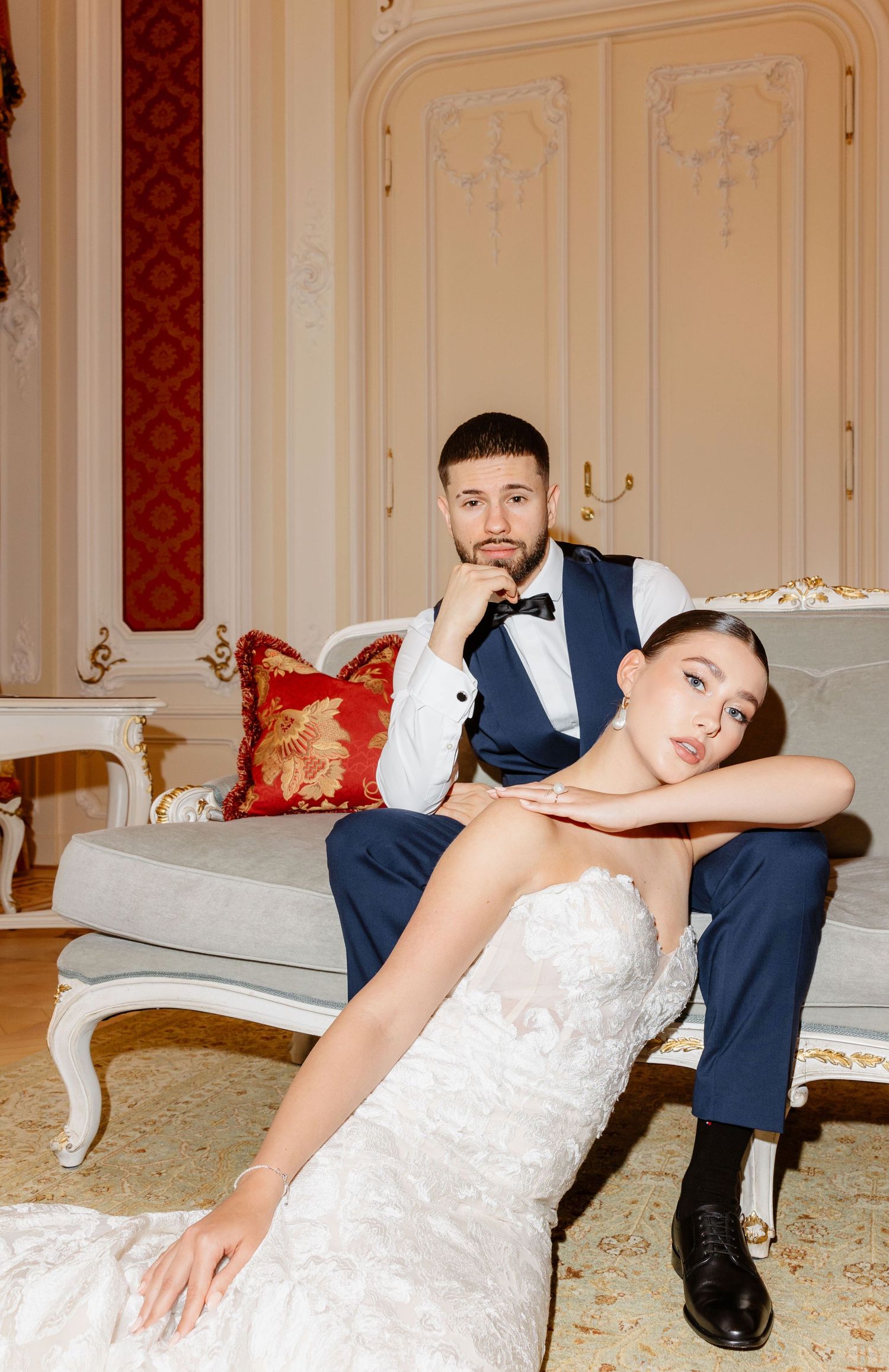 Bride in lace gown resting on floor while groom in navy suit sits on elegant sofa behind her.