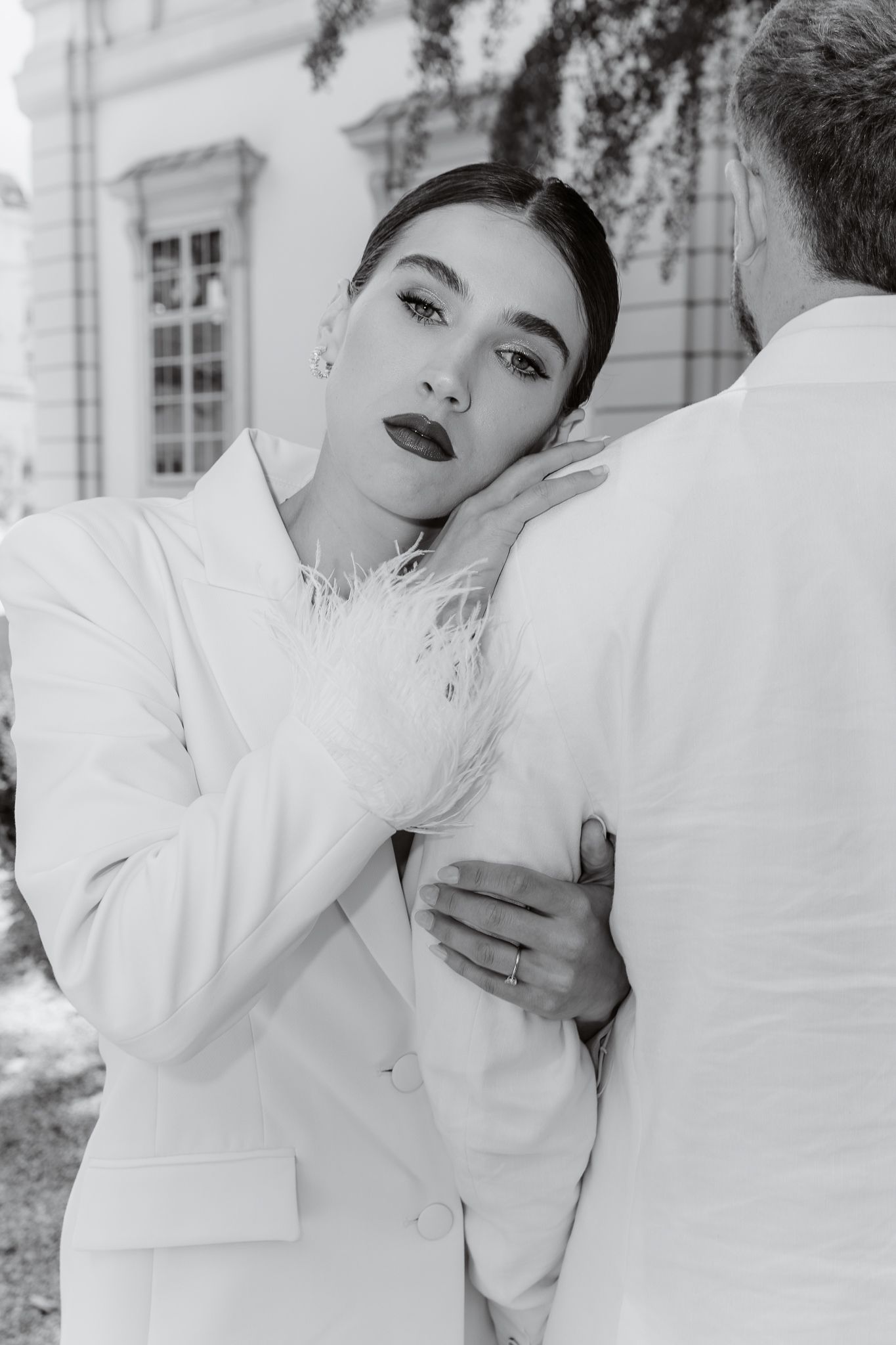 Bride in white feather blazer resting head on grooms shoulder in outdoor black and white photo.