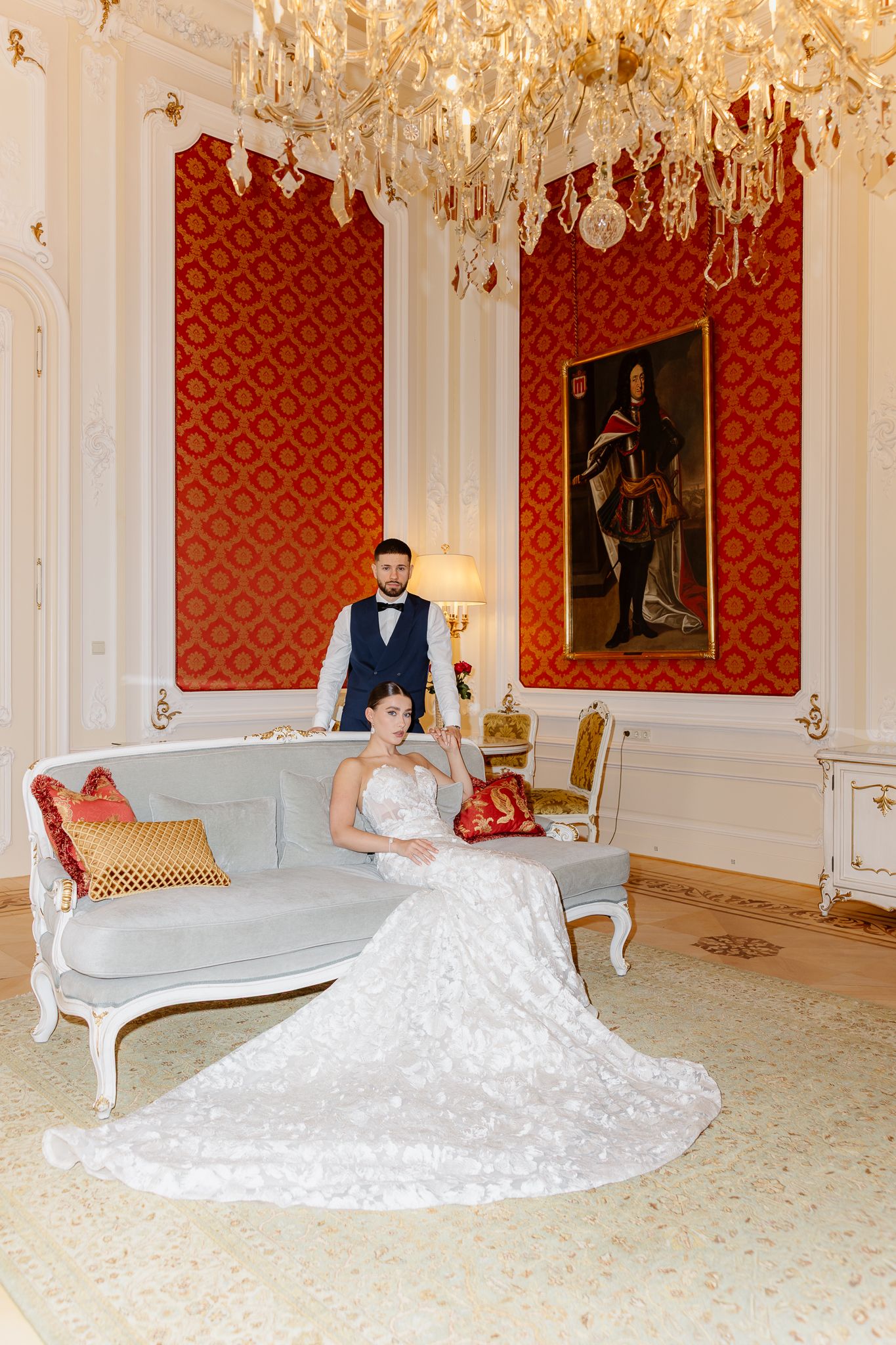 Bride and groom pose elegantly in ornate red and gold ballroom with crystal chandelier.