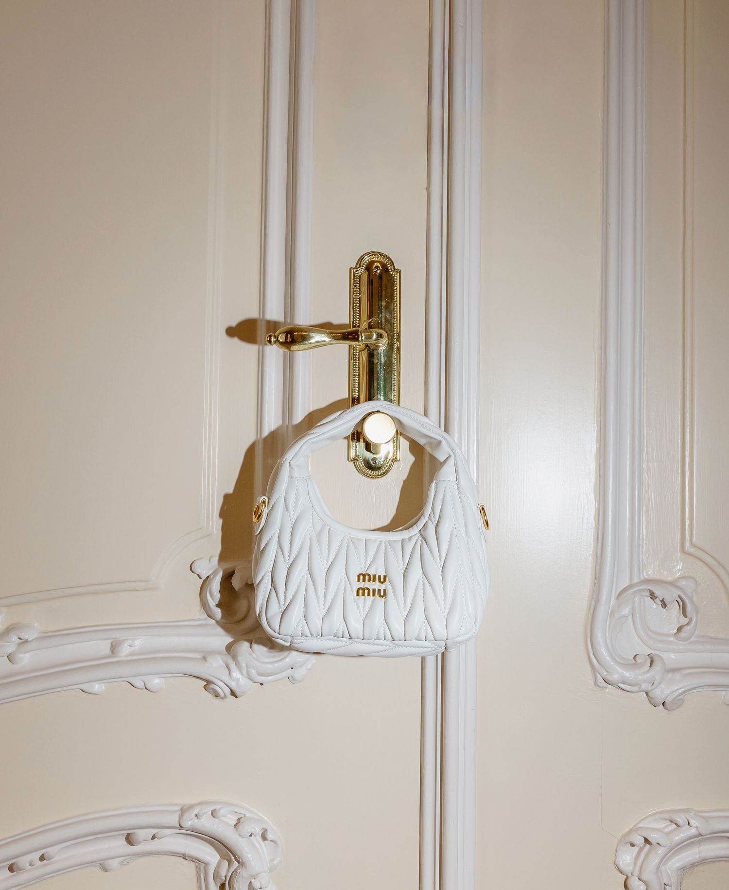 White quilted Miu Miu mini handbag with gold hardware hanging on ornate cream door handle.