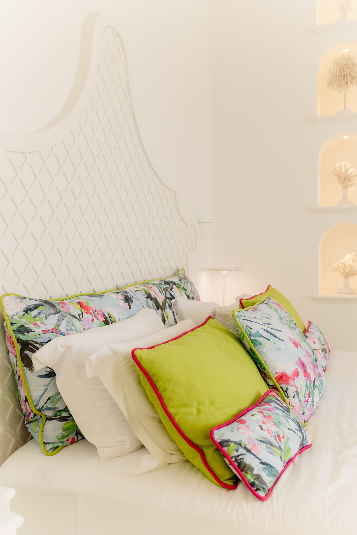 White bed with quilted headboard styled with green, floral, and white decorative pillows in bright room.