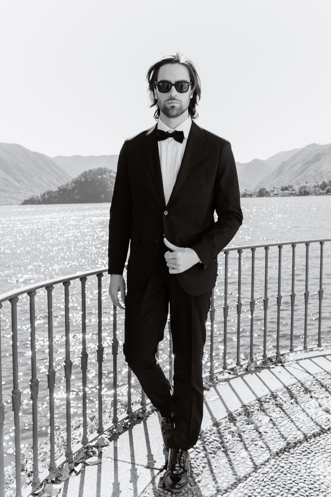 Elegant groom in black tuxedo and sunglasses posing by a waterfront railing with mountain backdrop.