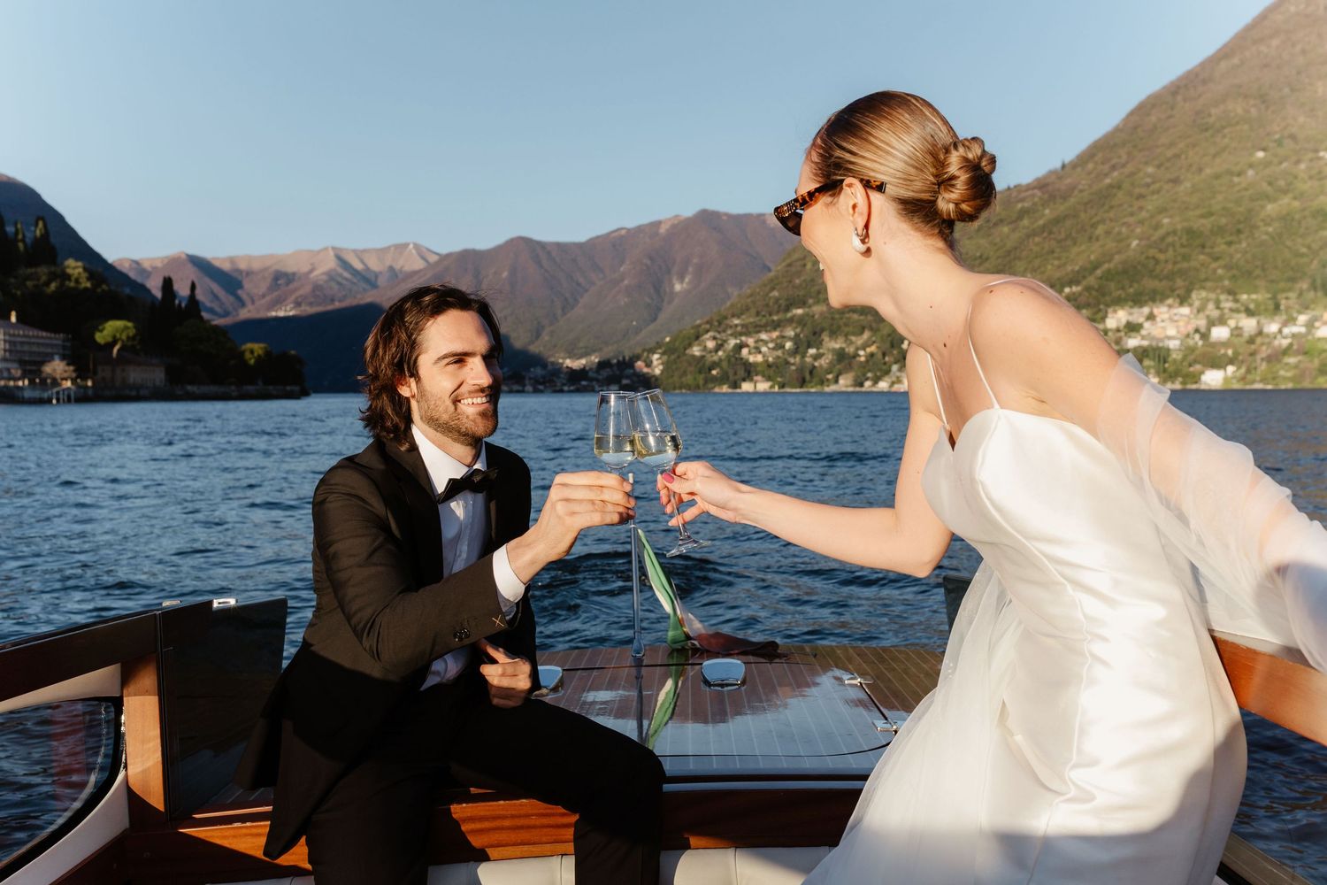 Bride and groom toasting champagne glasses on a wooden boat with scenic Italian lake and mountains behind them.
