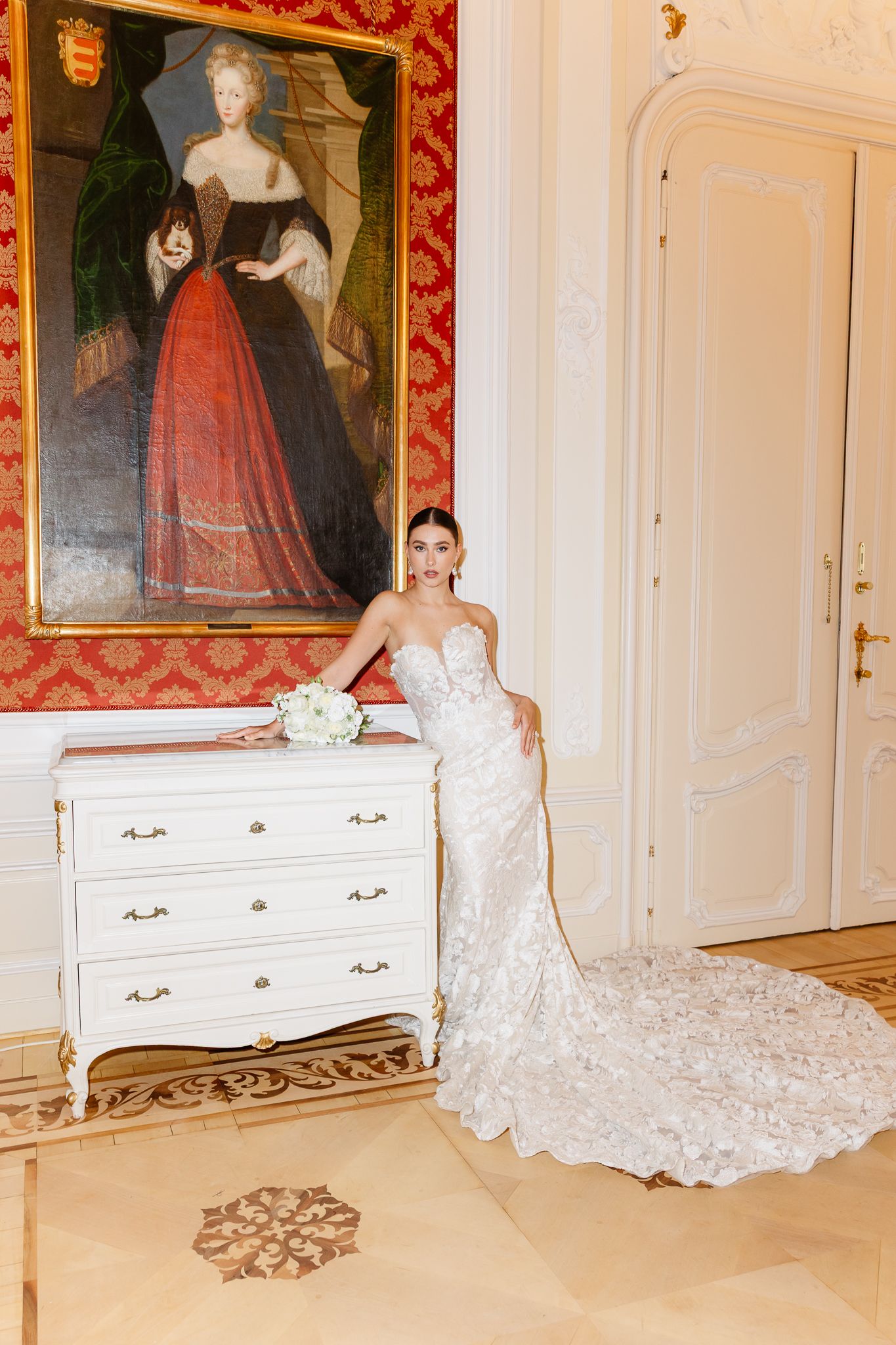 Elegant bride in lace mermaid dress poses beside white dresser beneath vintage portrait in opulent red room in Vienna,Austria.