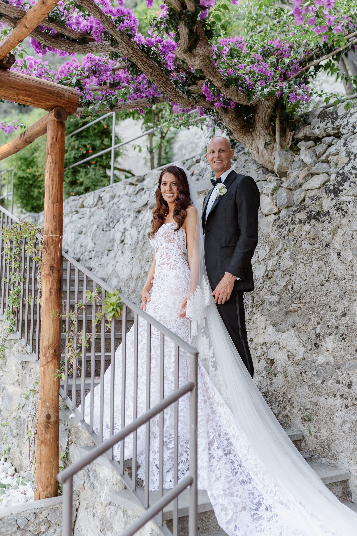 Bride and groom pose on stone staircase under purple bougainvillea, bride in lace gown with long train, groom in black tuxedo.
