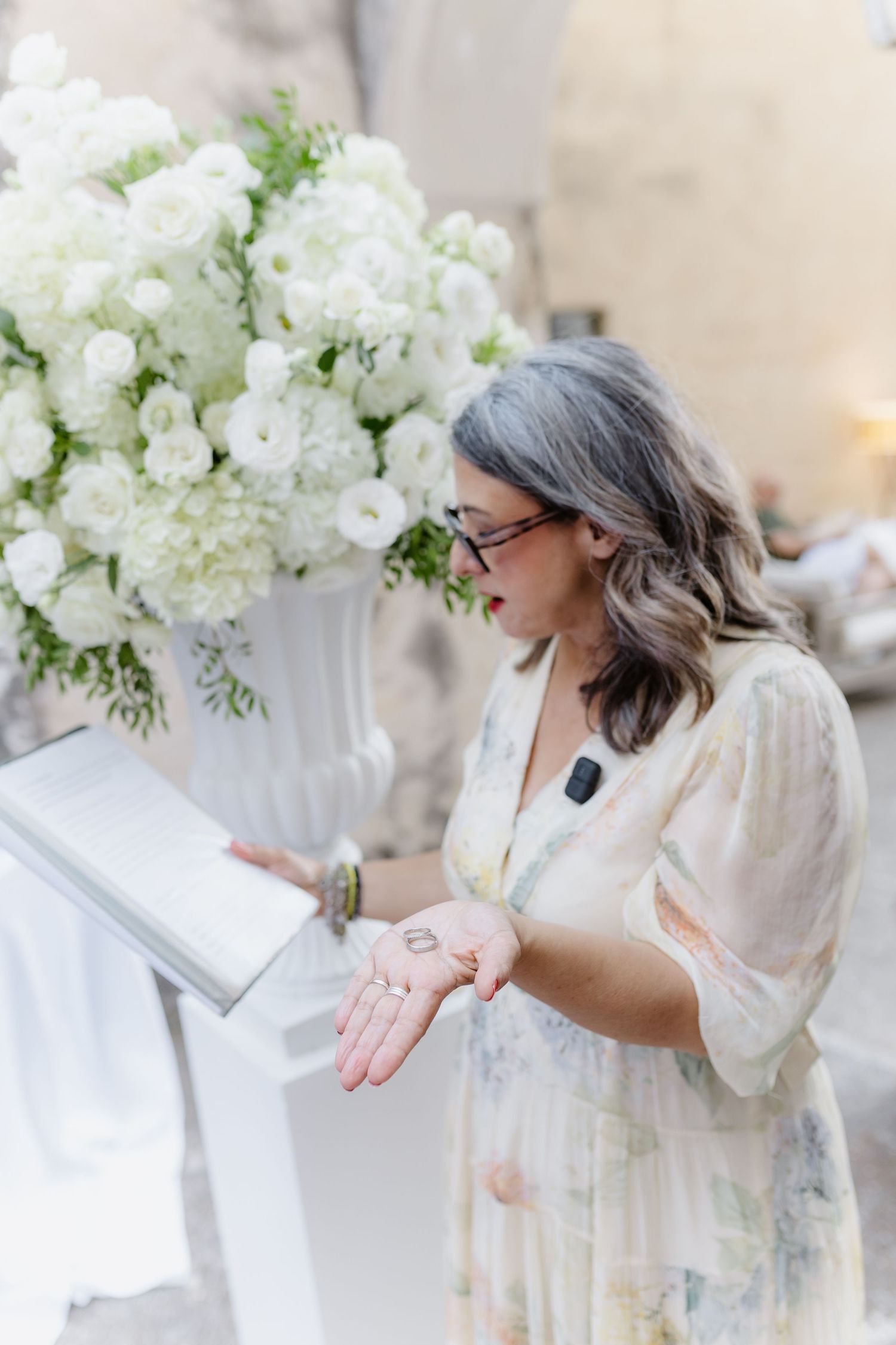 Woman in floral dress holding wedding rings at a ceremony podium with white flower arrangements.