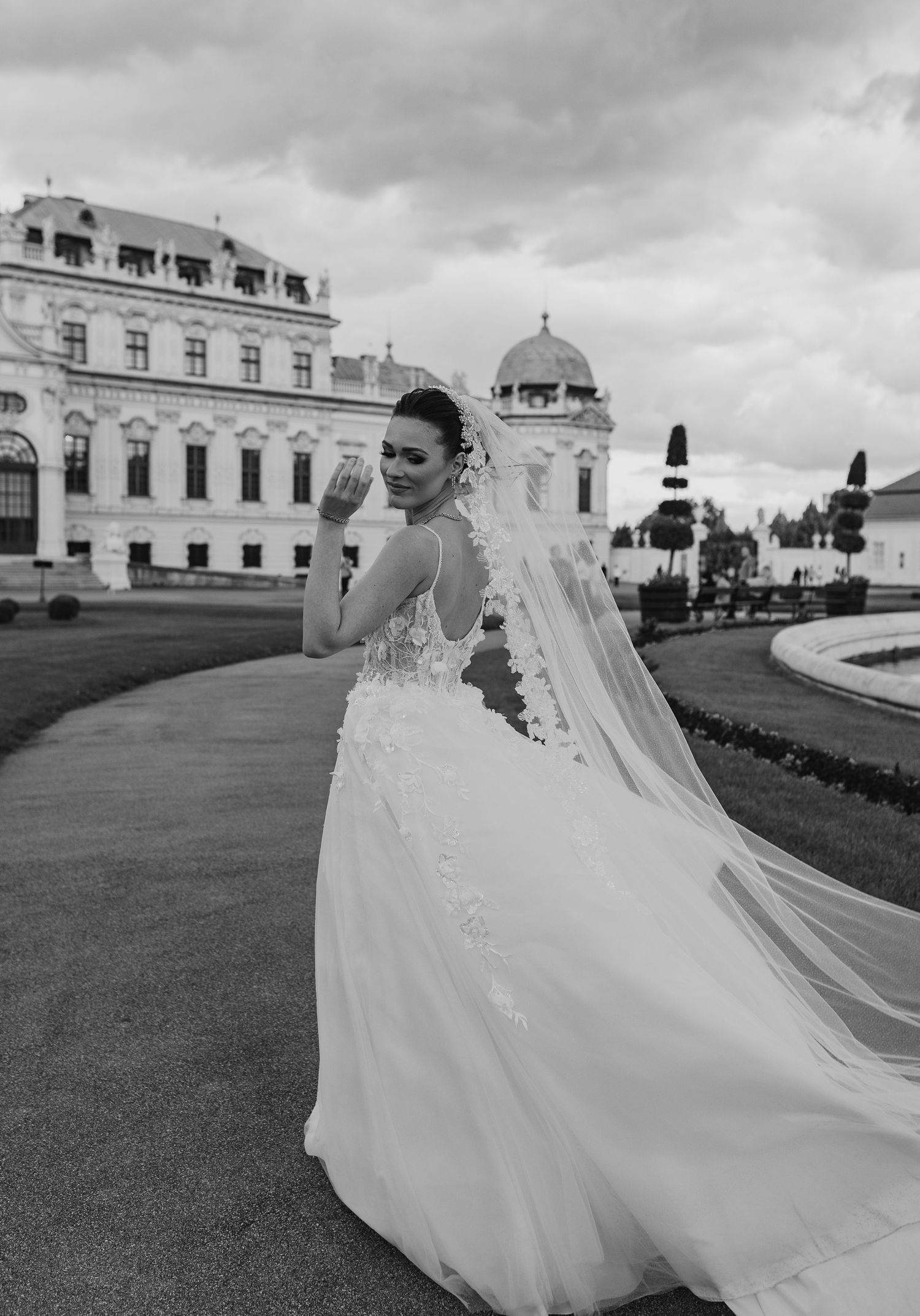 Smiling bride in lace wedding gown with flowing veil outside Belvedere Palace Vienna.