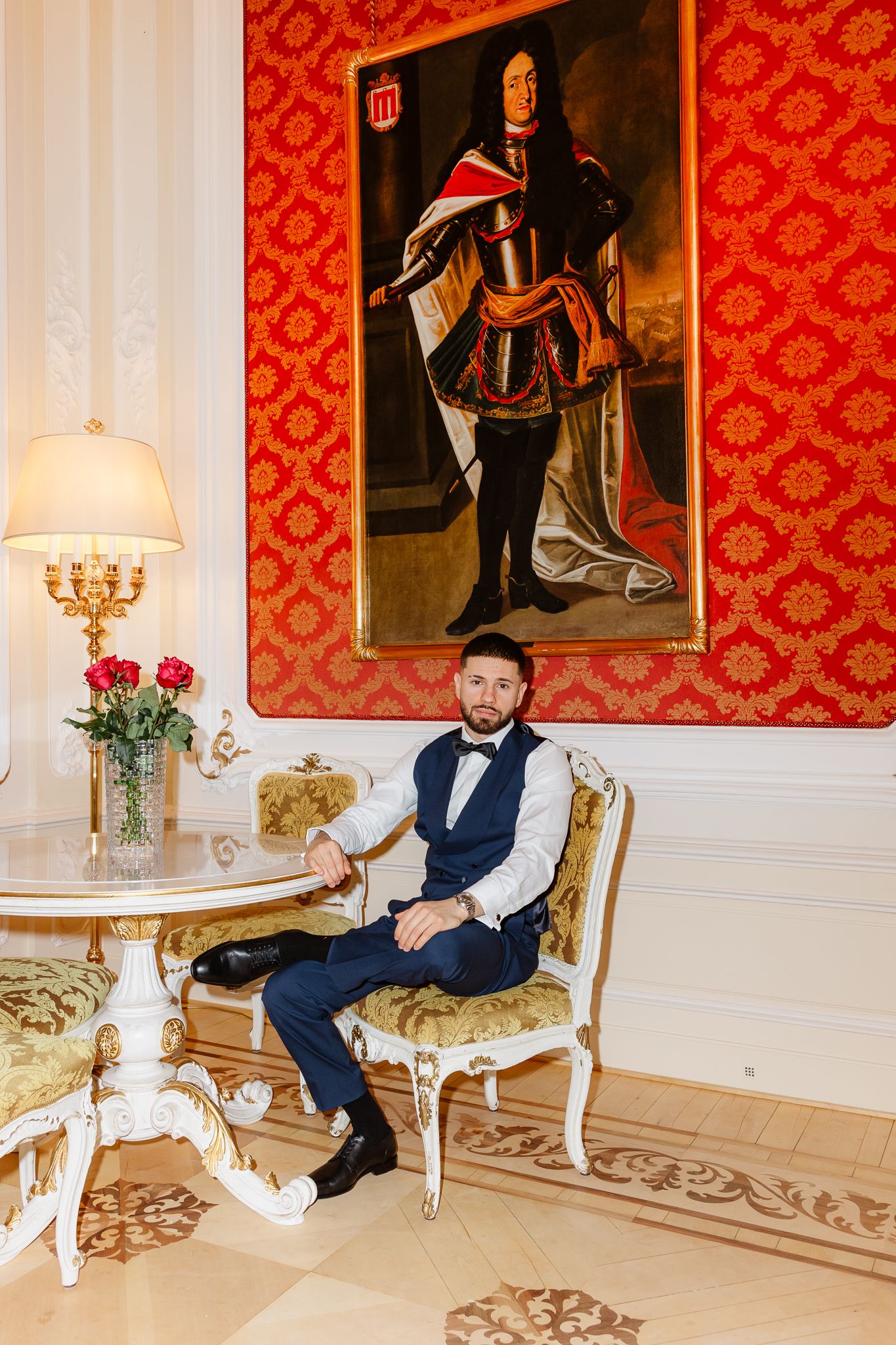 Groom in navy suit sits in gilded chair beneath large classical portrait in red damask luxury interior.