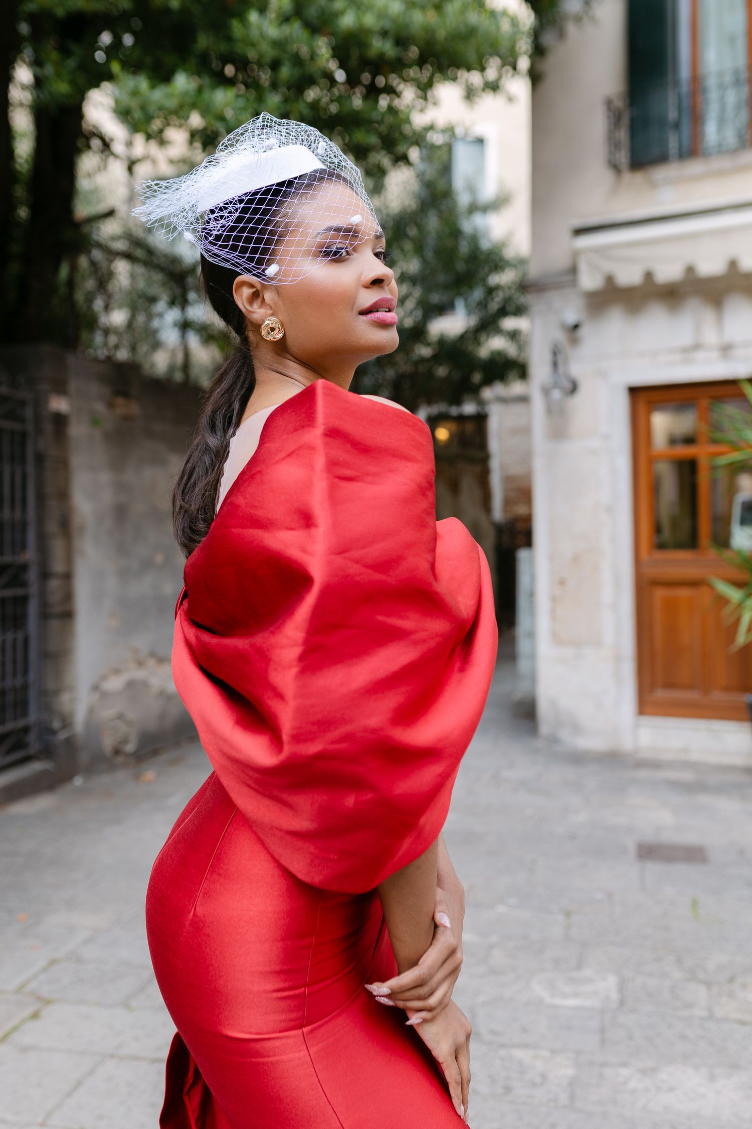 Elegant woman in red gown with dramatic sleeves and white fascinator hat poses on a European street.