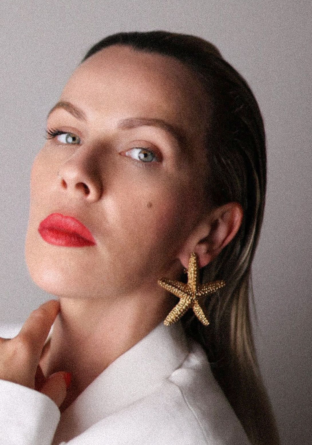 Close-up portrait of woman with blue eyes, red lips, and gold starfish earrings in white blazer.
