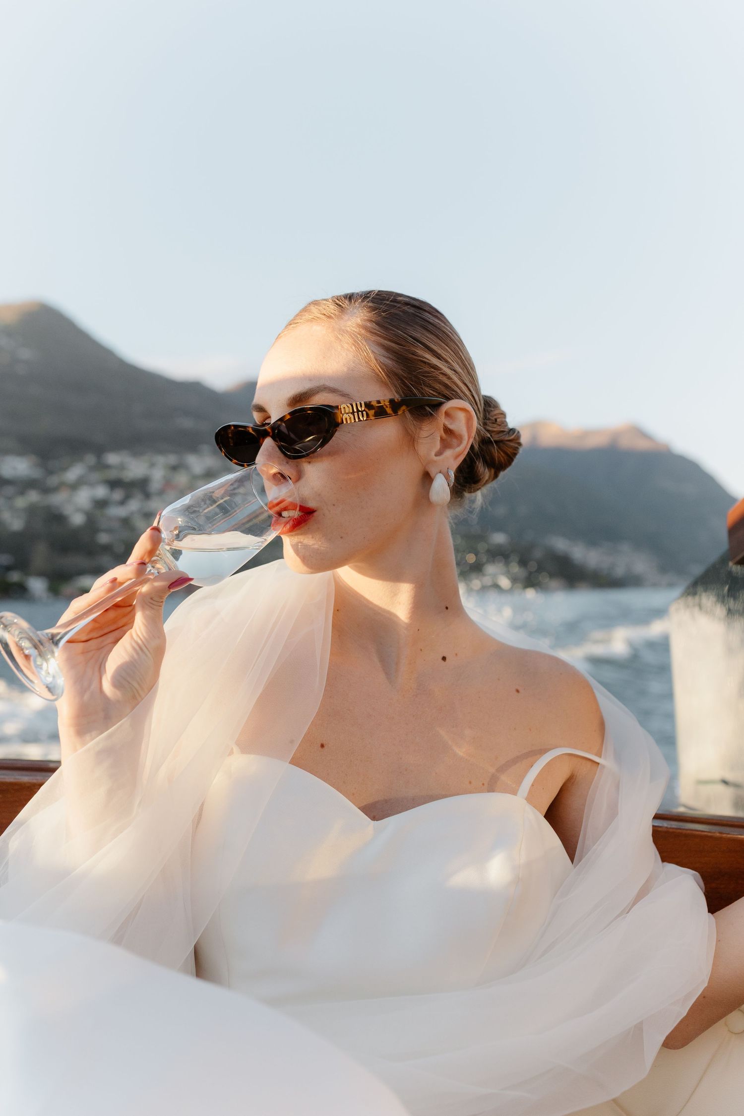 Elegant bride in white tulle gown and sunglasses sipping champagne on a luxury boat with mountain views.