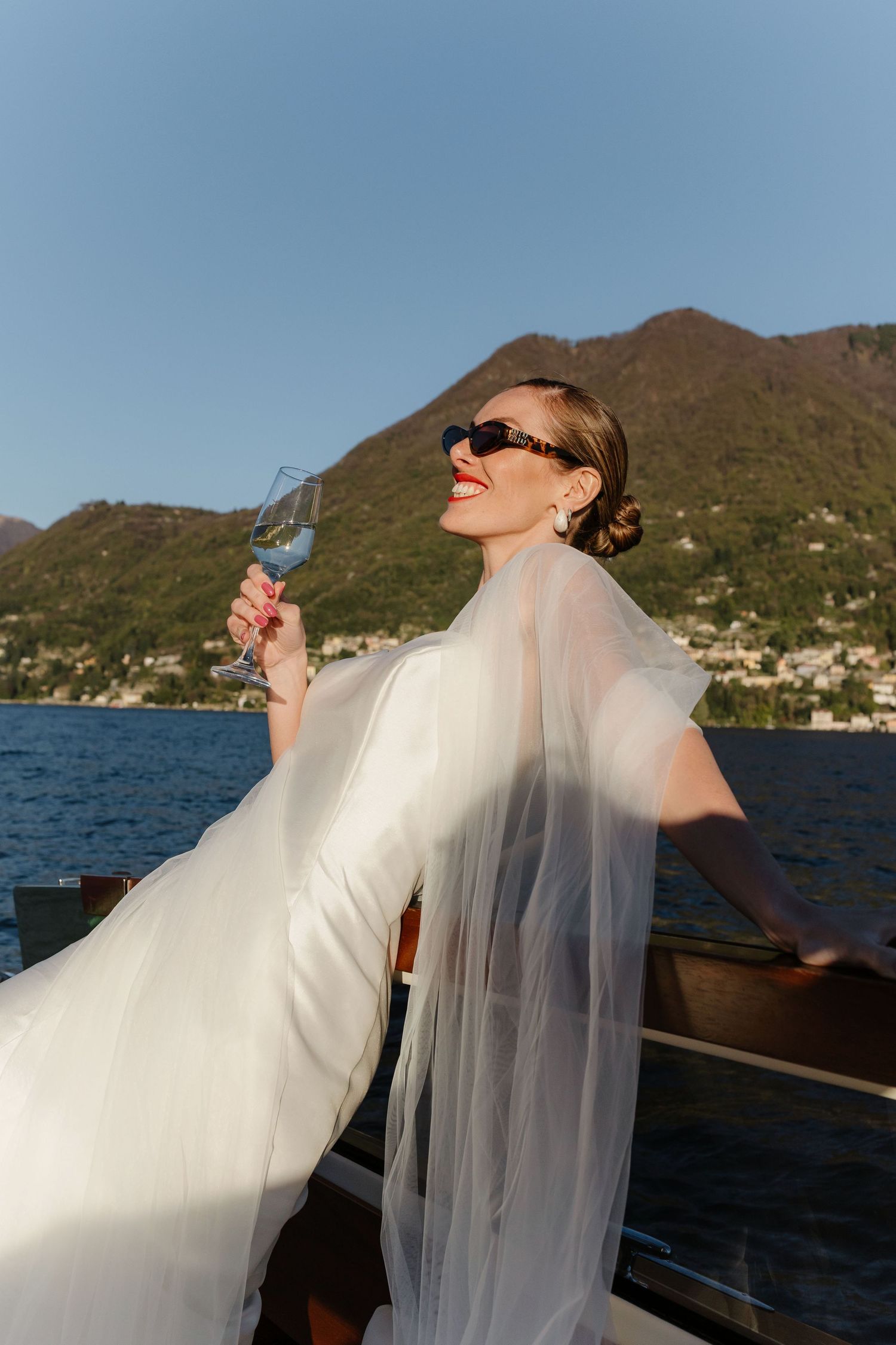 Elegant bride in white gown and sunglasses holding champagne glass on luxury boat with mountain lake backdrop.