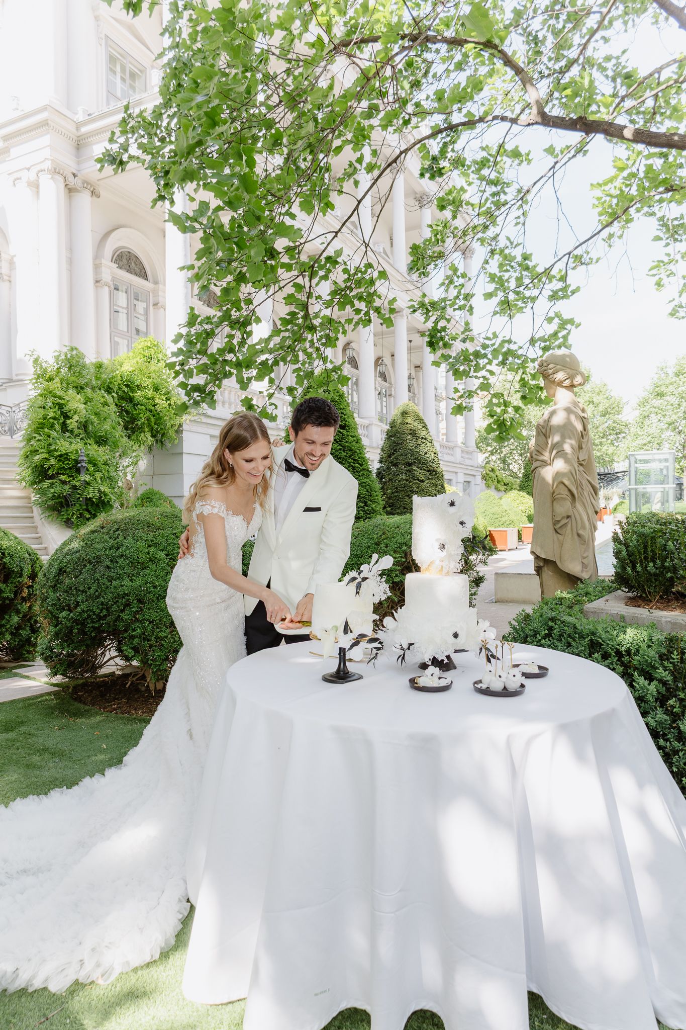 Bride and groom cutting wedding cake outdoors in elegant garden venue with white tiered cake.