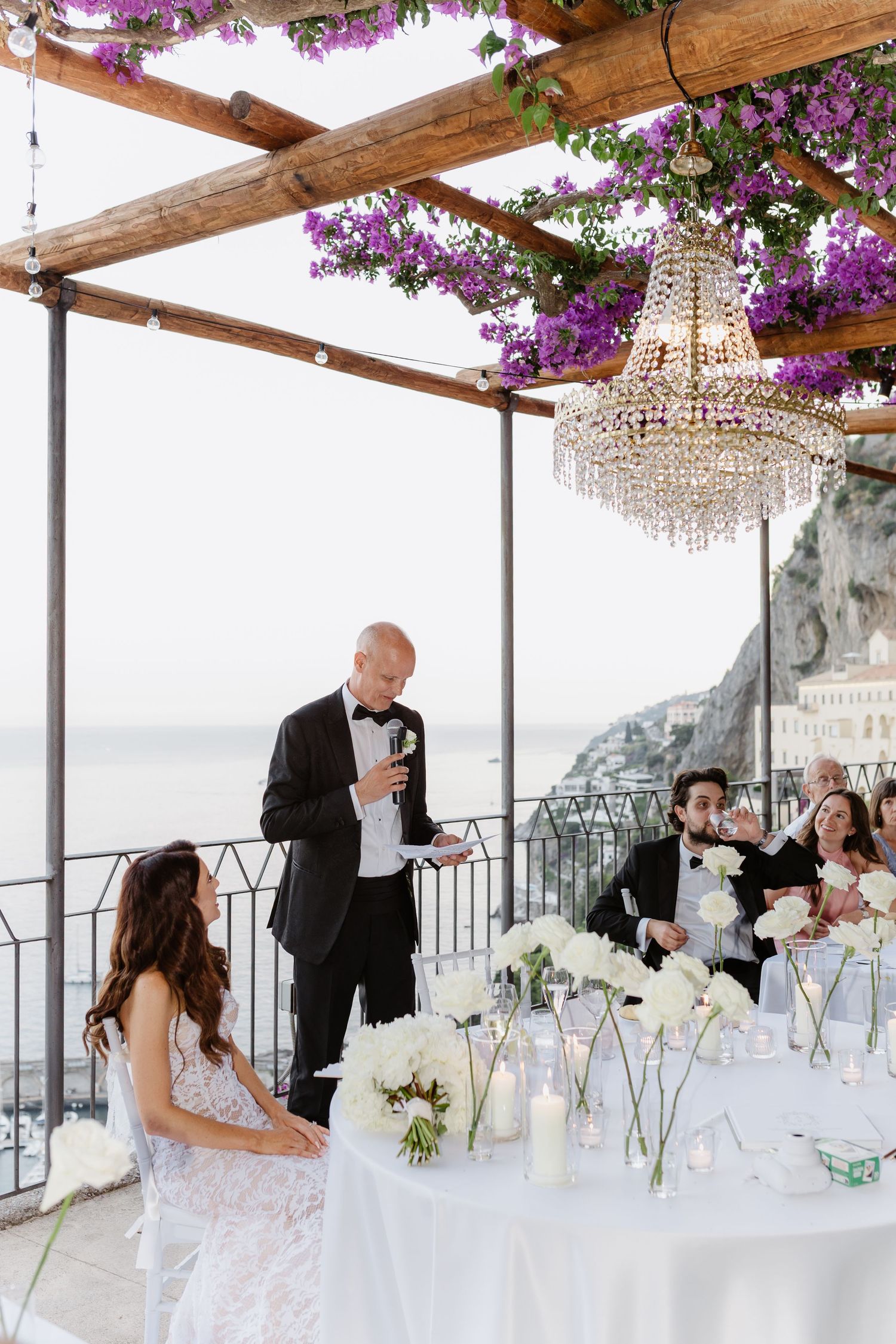 Elegant outdoor wedding reception on Amalfi Coast terrace with crystal chandelier, purple bougainvillea, and white floral centerpieces.