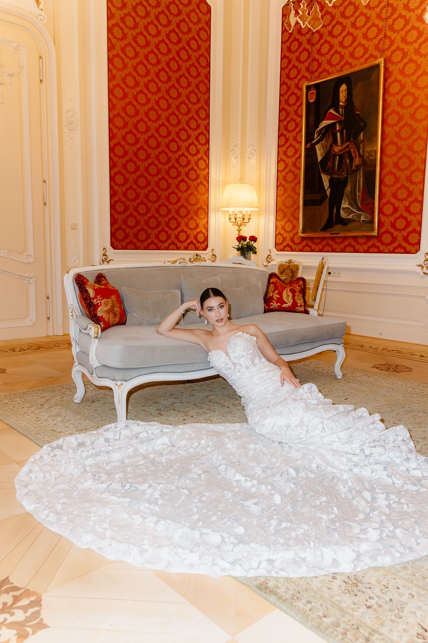 Bride in floral lace mermaid gown reclines on ornate sofa in luxurious red damask room with classic portrait.