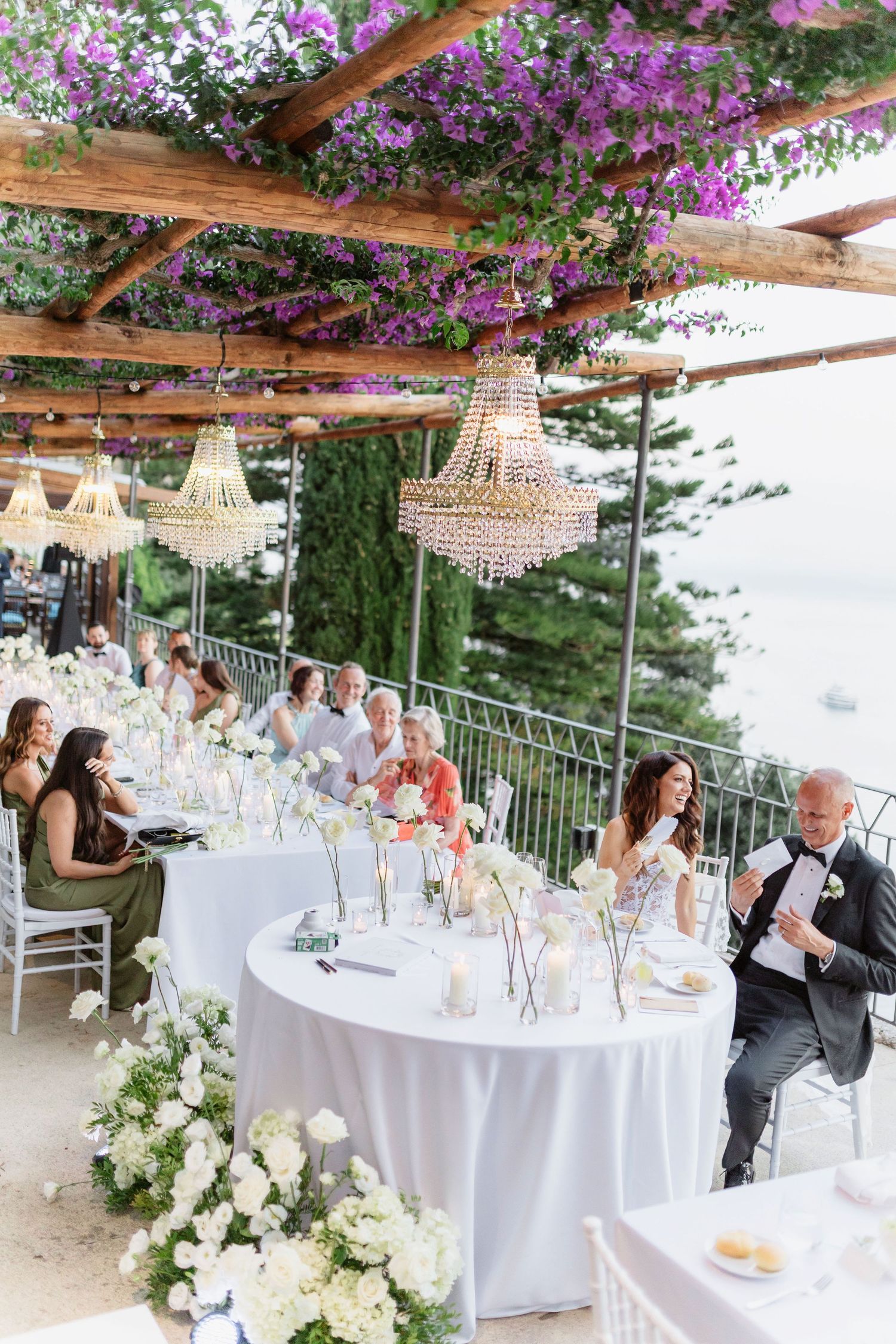 Elegant outdoor wedding reception with crystal chandeliers, white floral arrangements, and purple bougainvillea overhead near the sea.