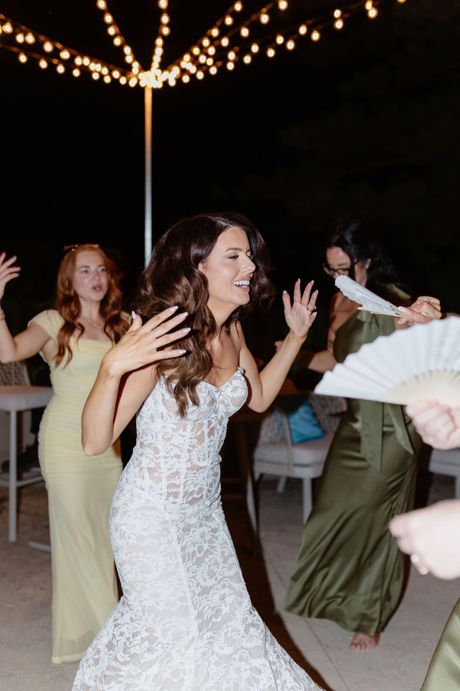 Bride in lace wedding gown dancing joyfully with bridesmaids in green dresses at outdoor evening reception.