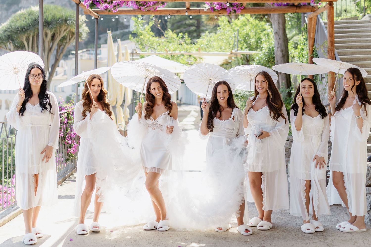 Bride and bridesmaids in white dresses and robes holding parasols posing outdoors at a sunny wedding venue.