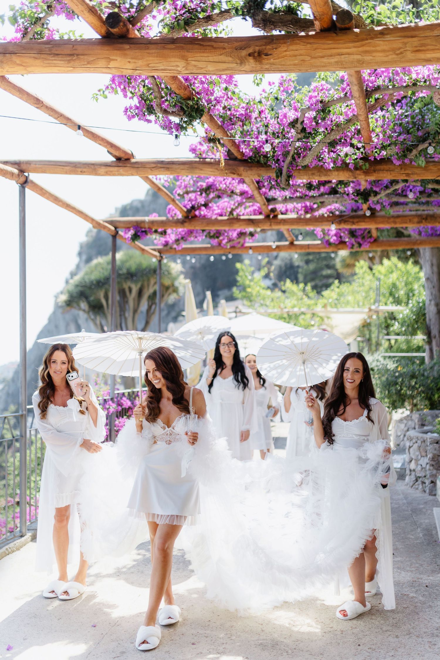 Bride and bridesmaids in white dresses holding parasols under a purple bougainvillea pergola on the Amalfi Coast.