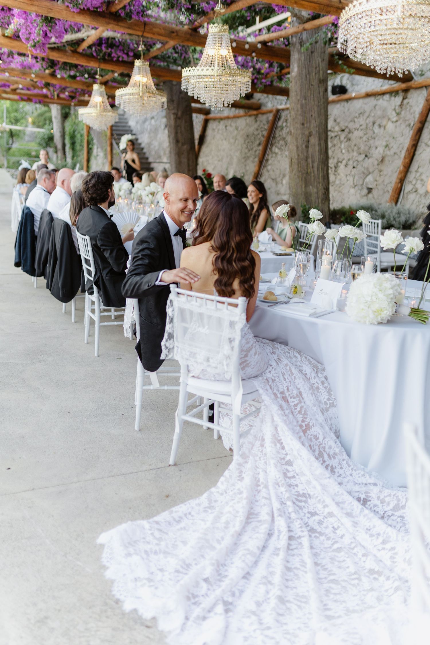 Bride in lace gown seated at elegant outdoor wedding reception with chandeliers and purple floral decor.