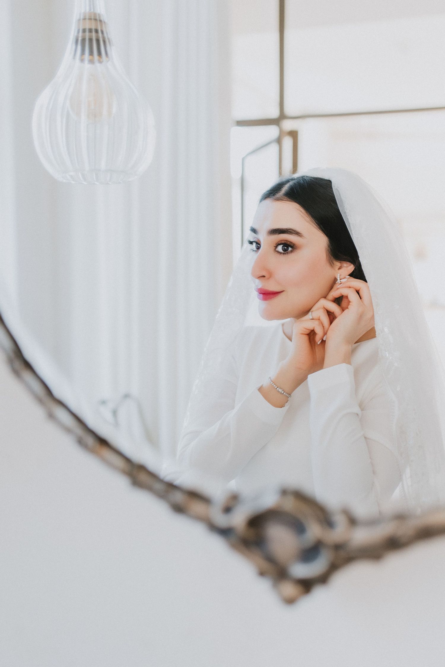 Elegant bride adjusting earring while gazing into an ornate mirror in soft natural light.