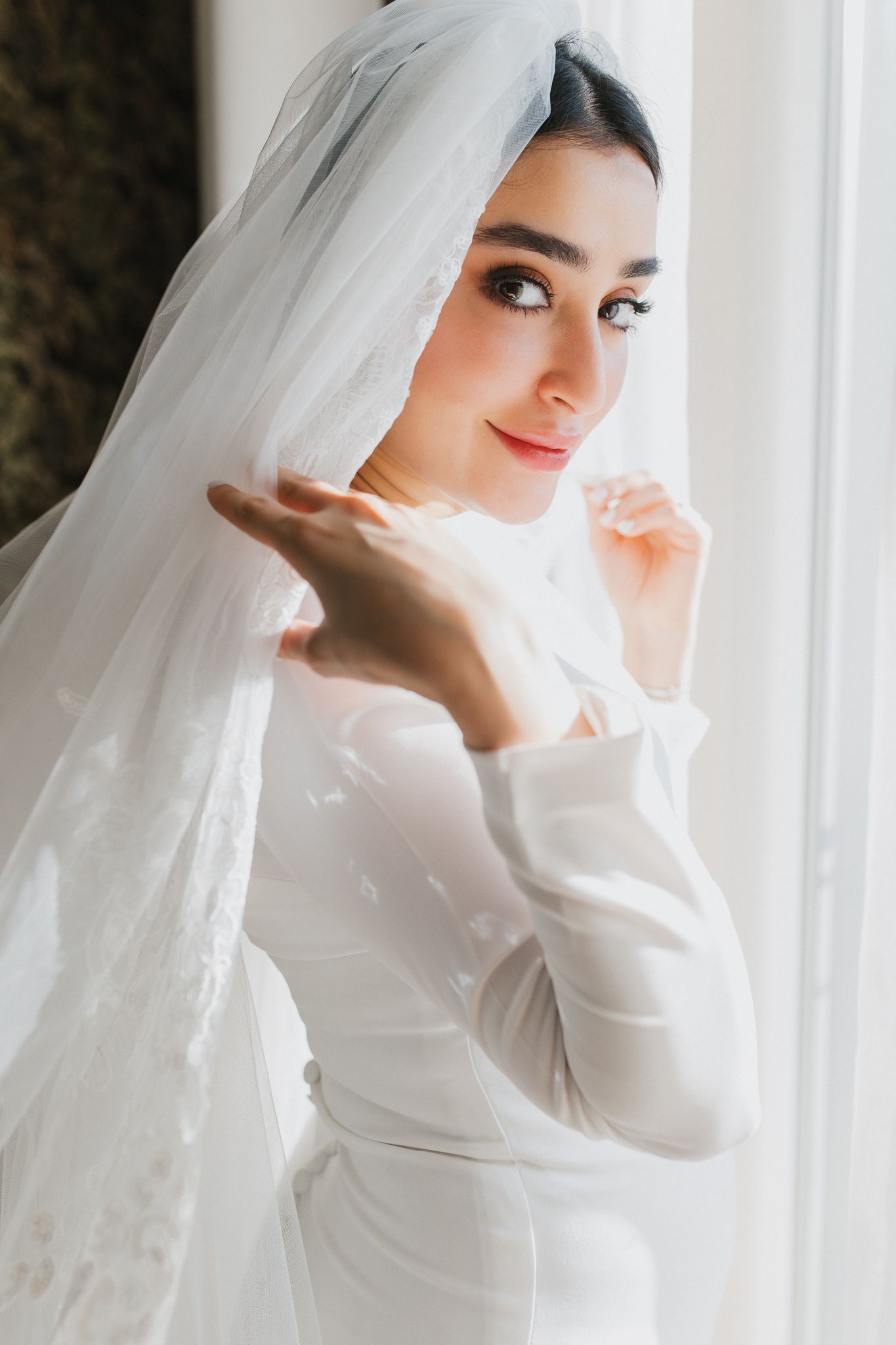Beautiful bride in white long-sleeve gown and veil posing gracefully by a bright window.