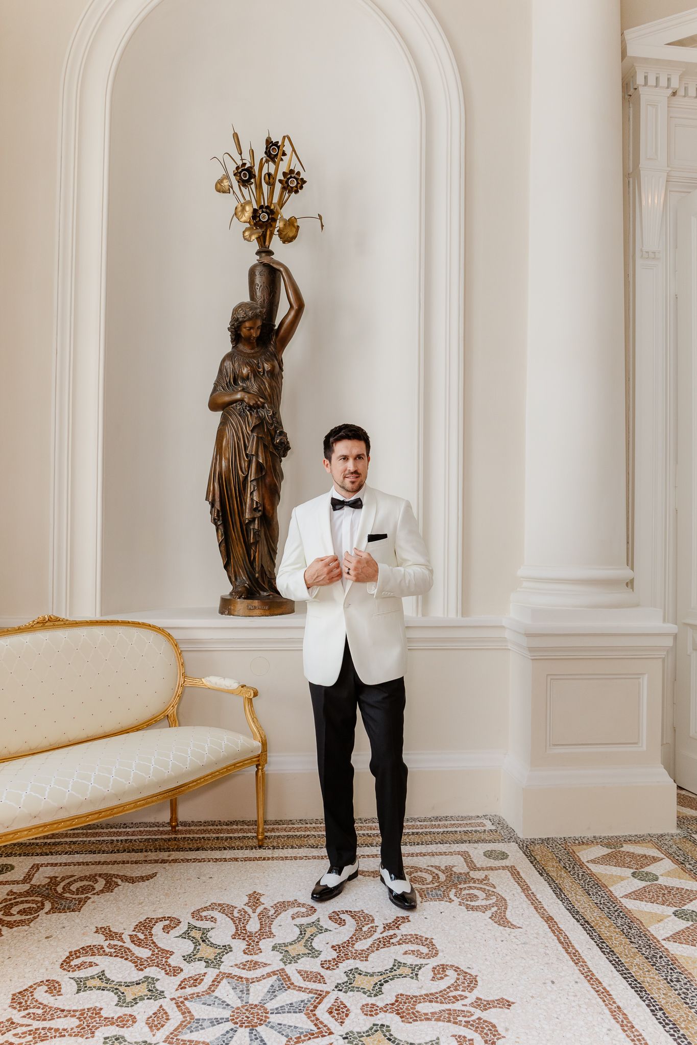 Dapper groom in white tuxedo jacket and black trousers posing in ornate classical interior.