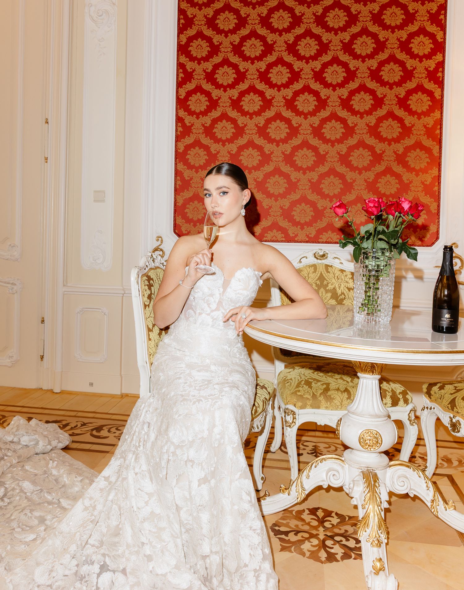 Elegant bride in lace gown holds champagne while seated at an ornate table in a red-walled room.