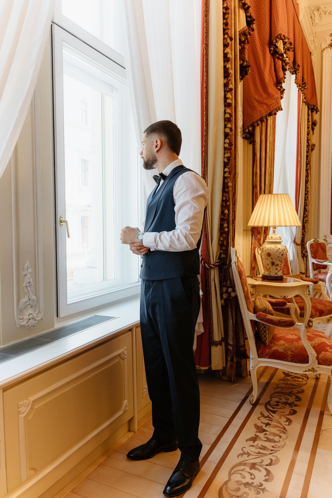Groom in navy vest and bow tie standing at window holding coffee cup in opulent hotel room.