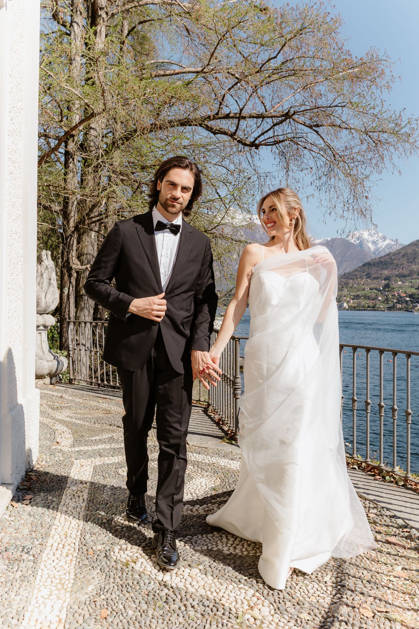 Elegant couple walks hand in hand along scenic lakeside promenade on their wedding day.