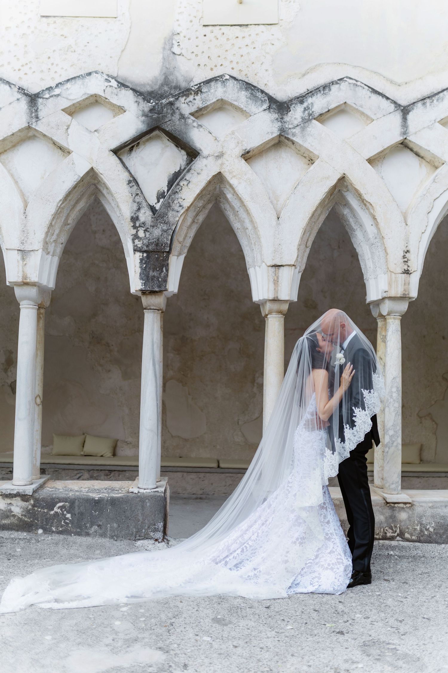 Bride and groom kissing under a flowing lace veil beside Gothic arches at an elegant wedding venue.