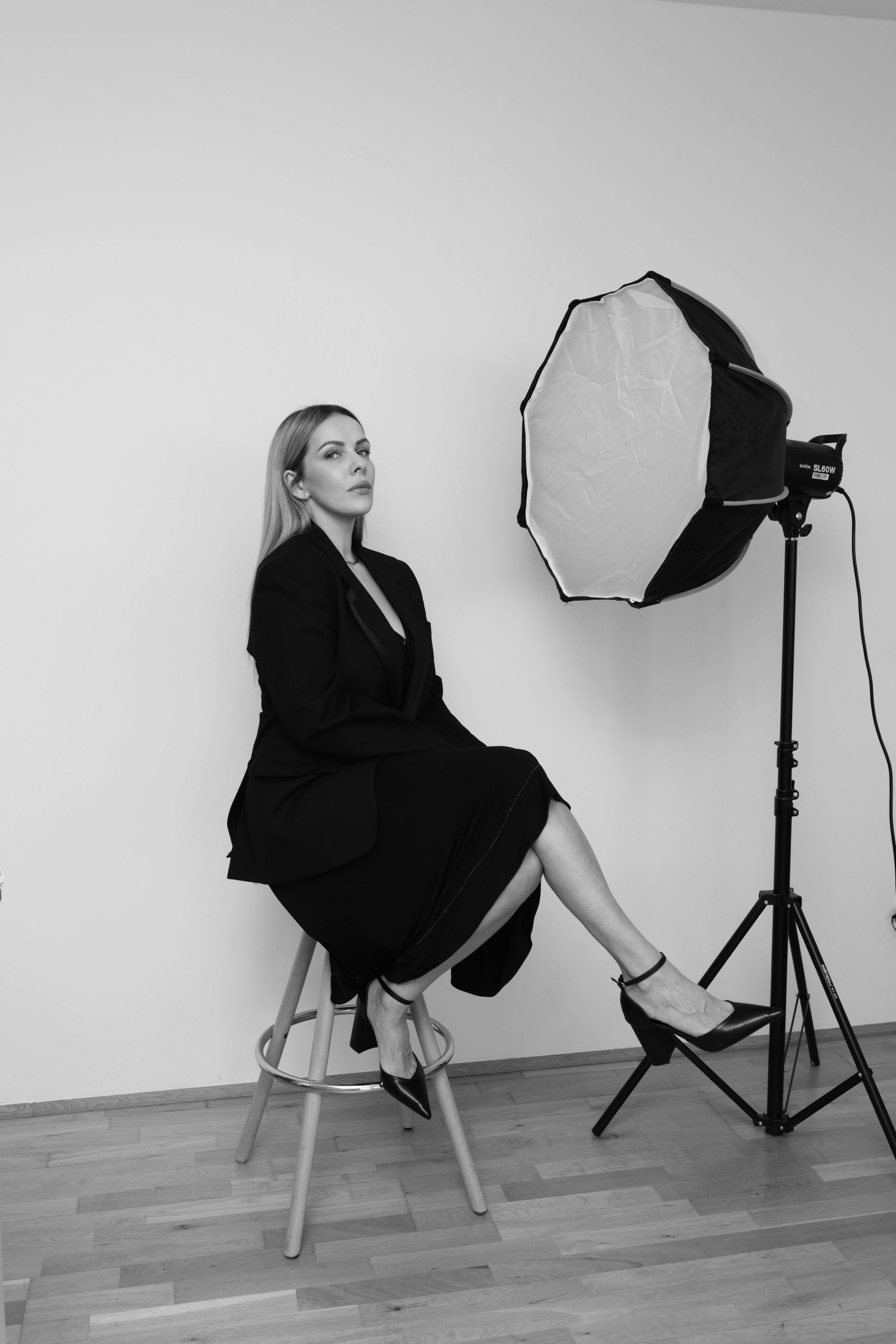 Black and white portrait of blonde woman in black dress seated on stool next to studio softbox light.