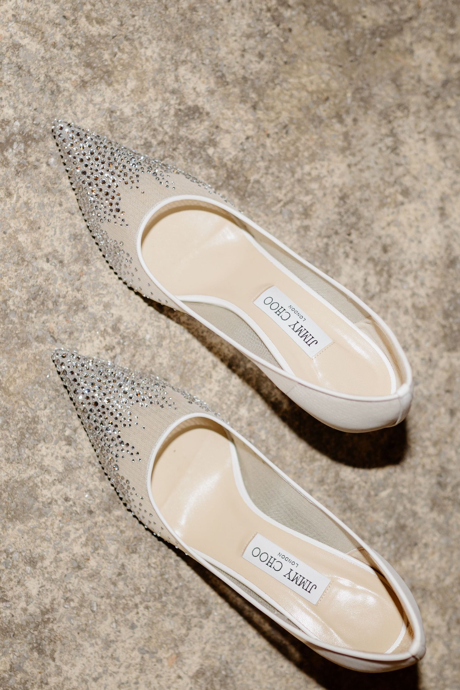 Jimmy Choo crystal-embellished pointed-toe ivory satin pumps displayed on a stone surface.
