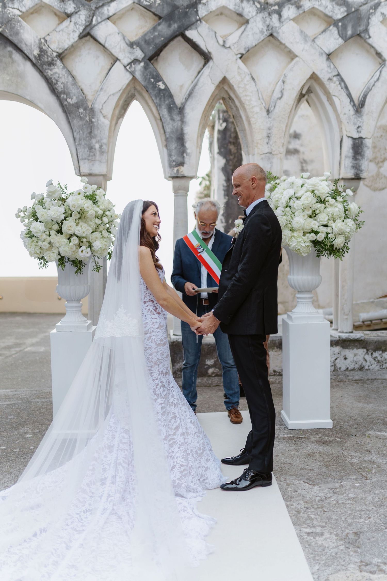Bride and groom exchange vows at an elegant outdoor wedding ceremony with white floral arrangements and Gothic stone arches.