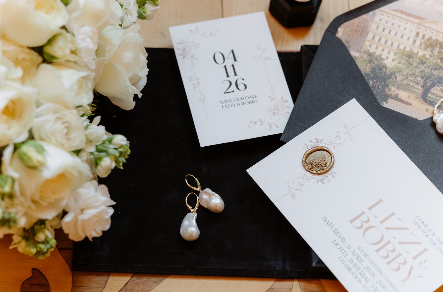 Elegant wedding stationery flat lay with white roses, pearl drop earrings, and save the date card.