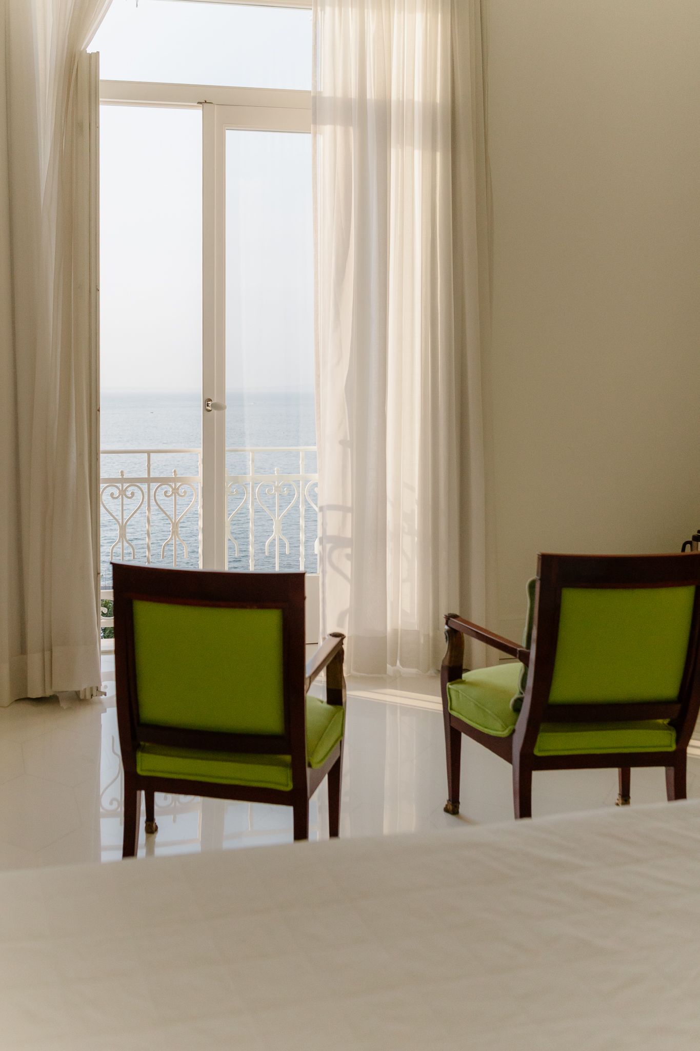 Two green wooden chairs facing open balcony doors with ocean view in a bright white room.