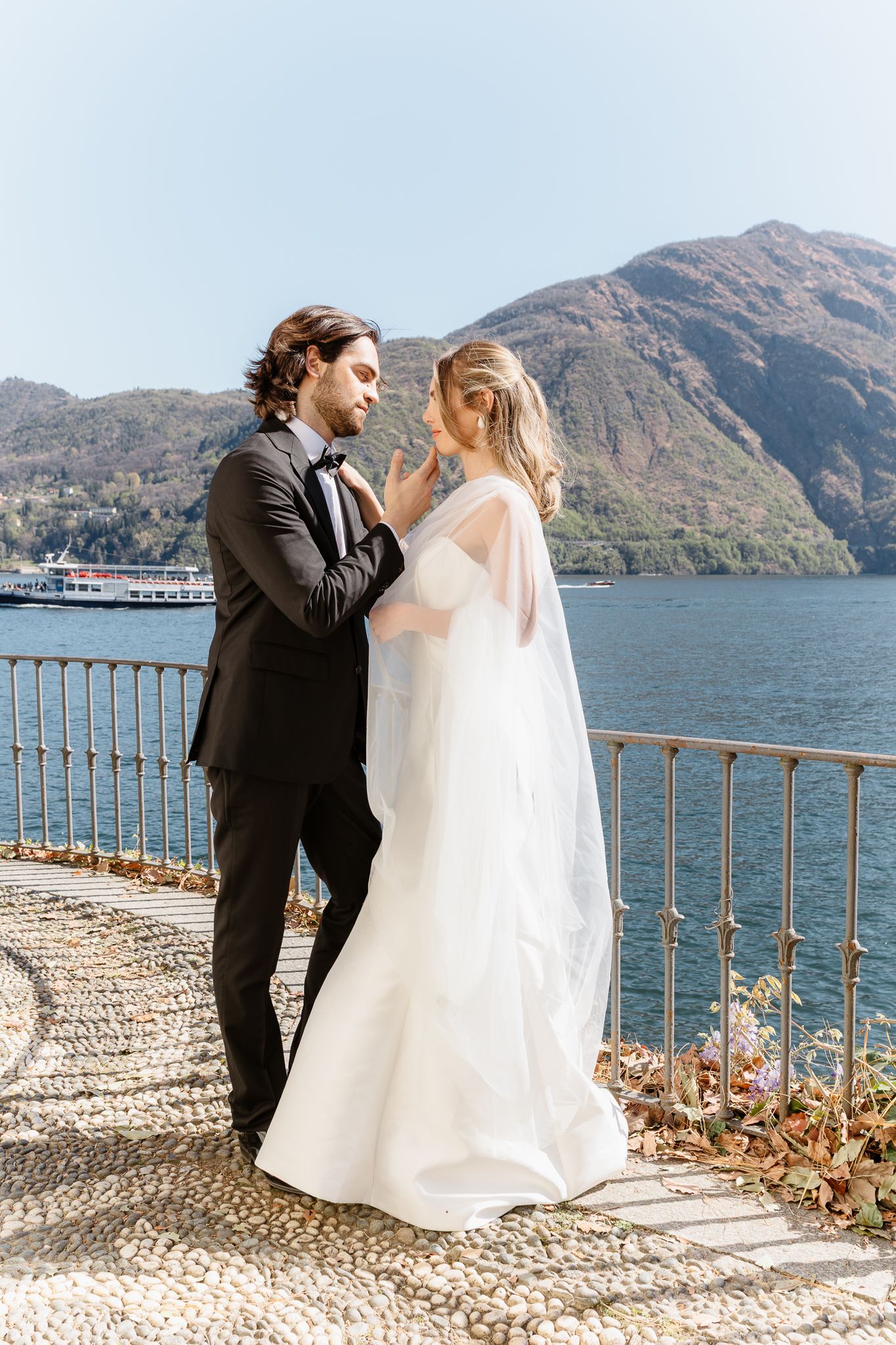 Bride and groom share romantic moment on lakeside terrace with mountain backdrop on wedding day.