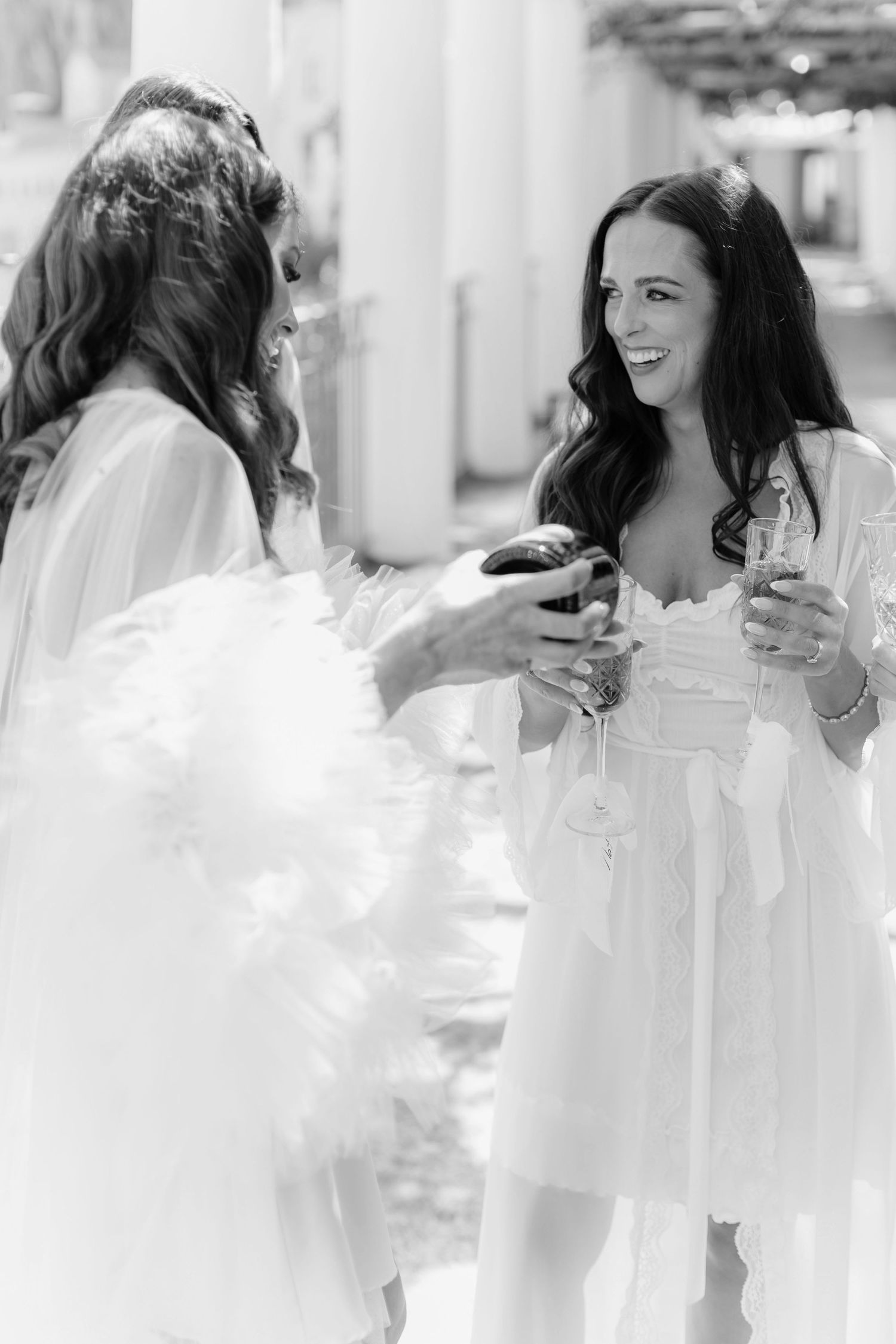 Two women in white robes laughing and holding champagne glasses during a bridal party celebration.