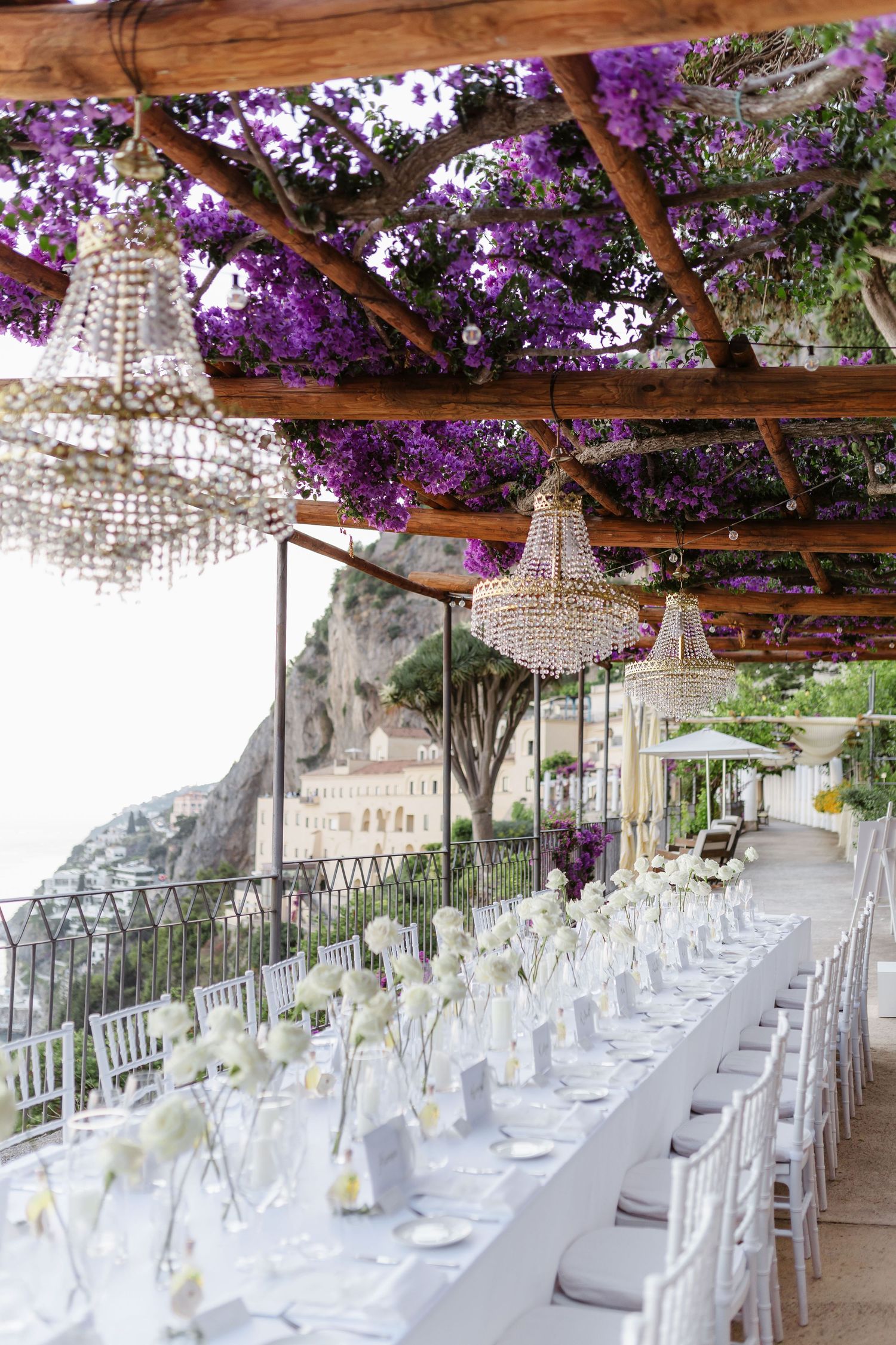 Elegant outdoor wedding reception under a pergola with crystal chandeliers, purple bougainvillea, and long white-draped tables.