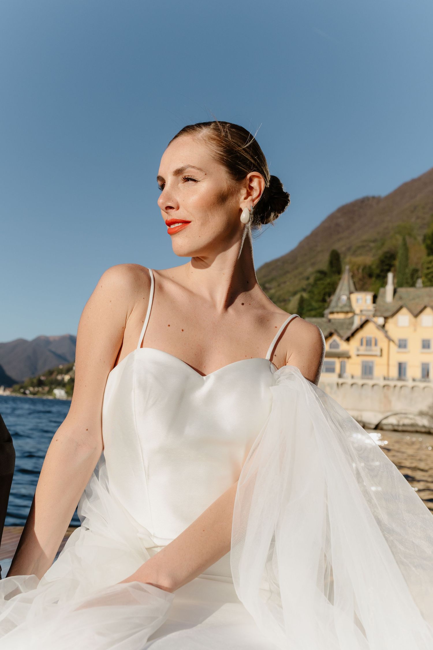 Radiant bride in white satin gown with tulle sitting by a sunlit Italian lake with mountain backdrop.